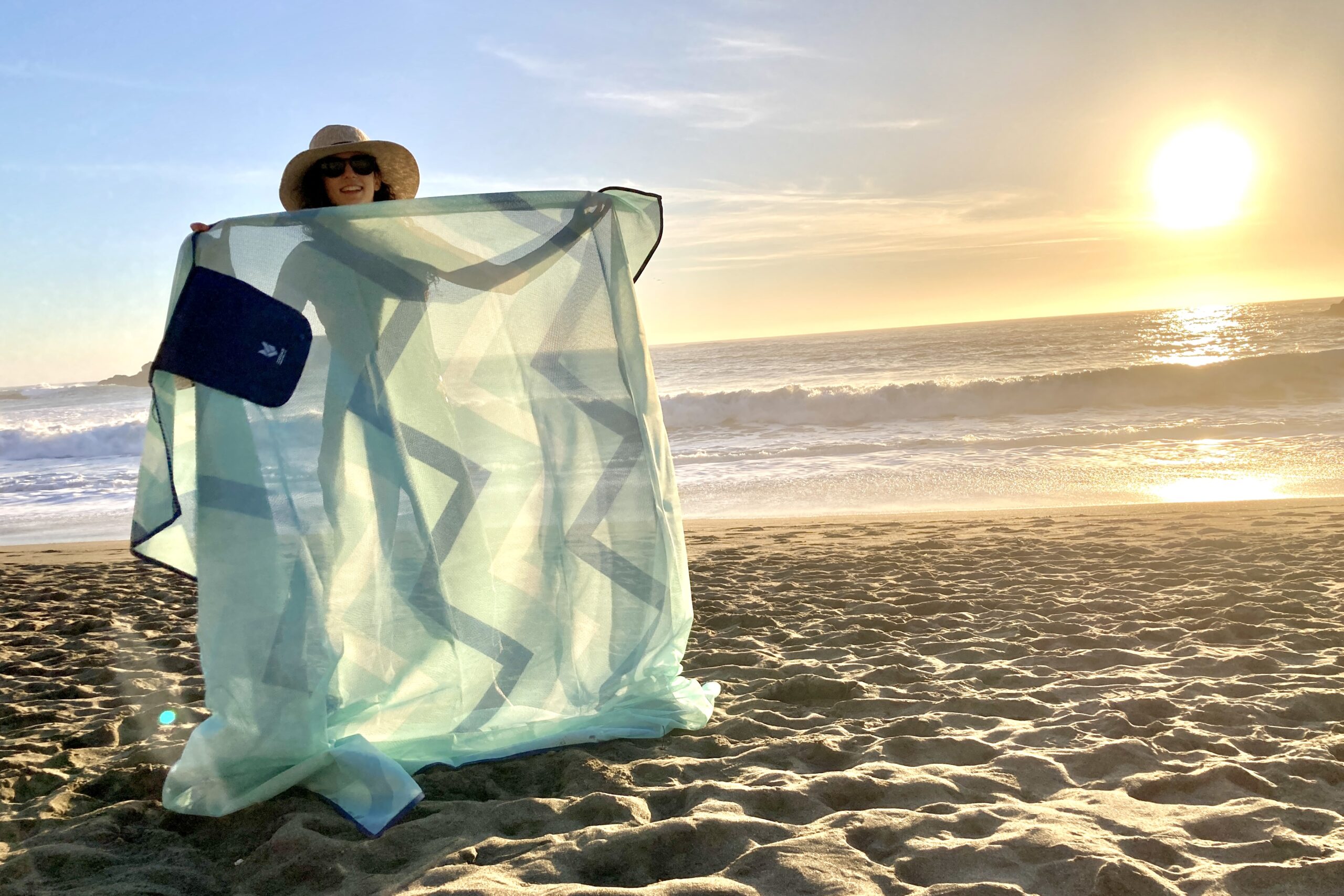 Person holding up the CGear Multimats Sandlite Sand-free Mat at the beach in the light to show its mesh and almost translucent material.