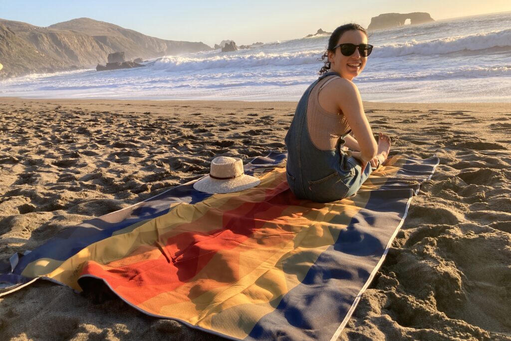 Woman sitting on the Nemo Victory Patio Blanket on the beach, looking over her shoulder at the camera.