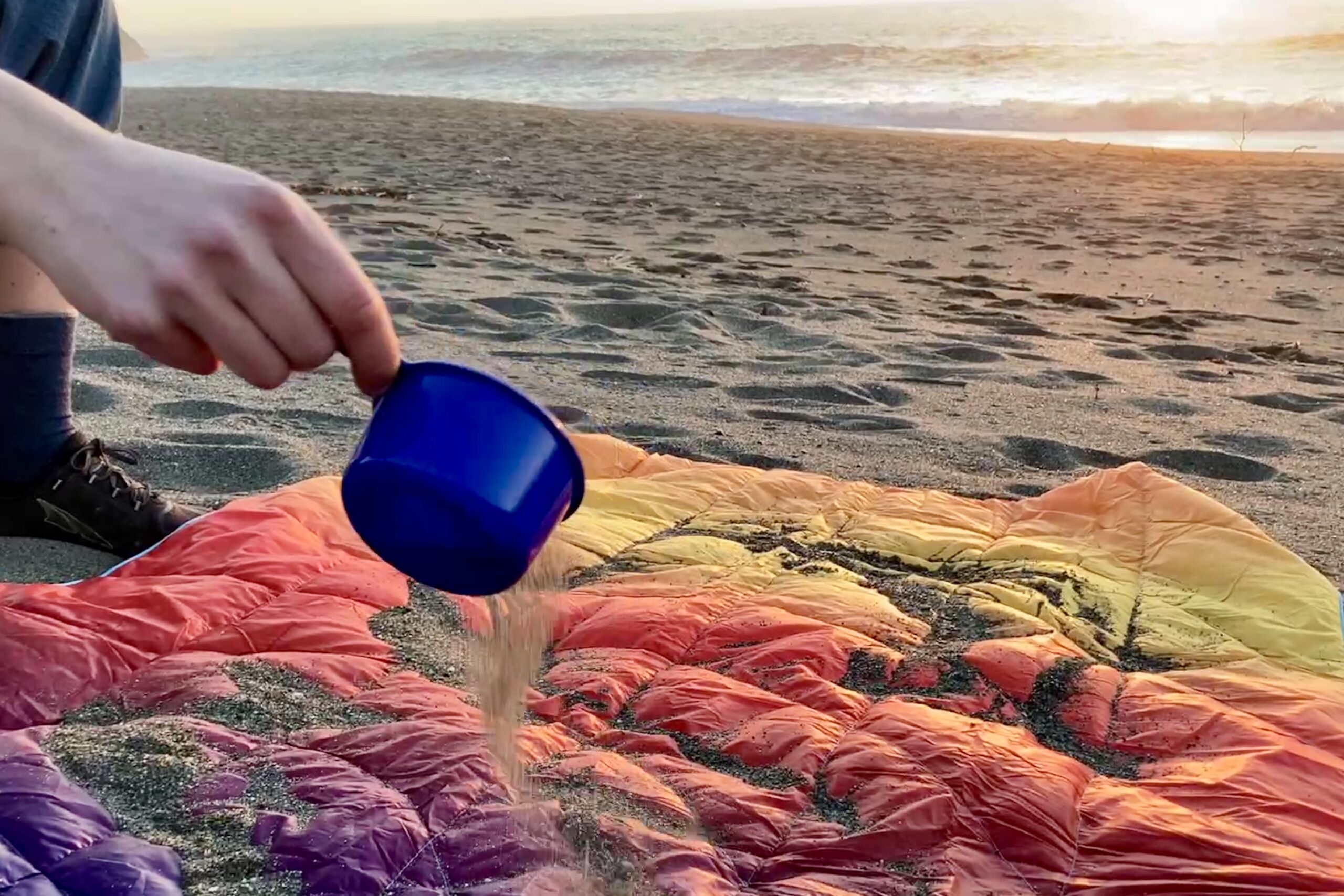 Person at the beach pouring sand over the Rumpl NanoLoft Puffy Blanket in preparation for a shake test.