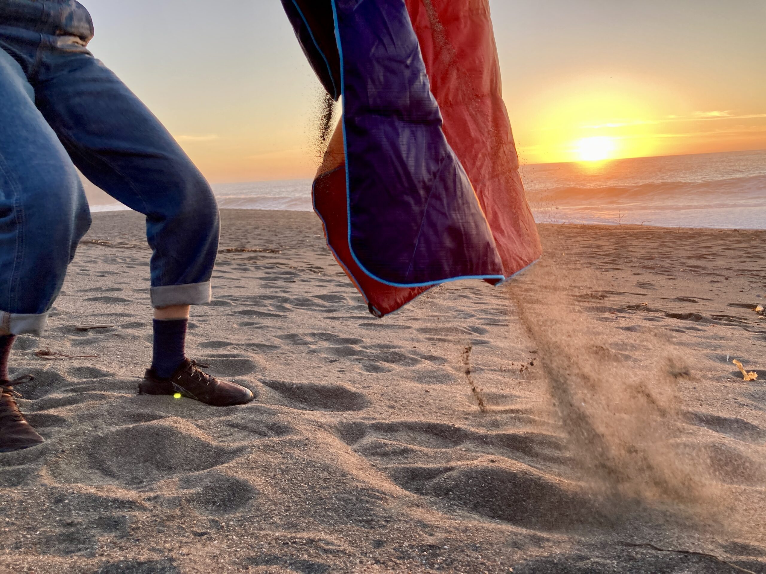 A person shaking sand off the Rumpl NanoLoft Puffy Blanket on the beach during sunset.