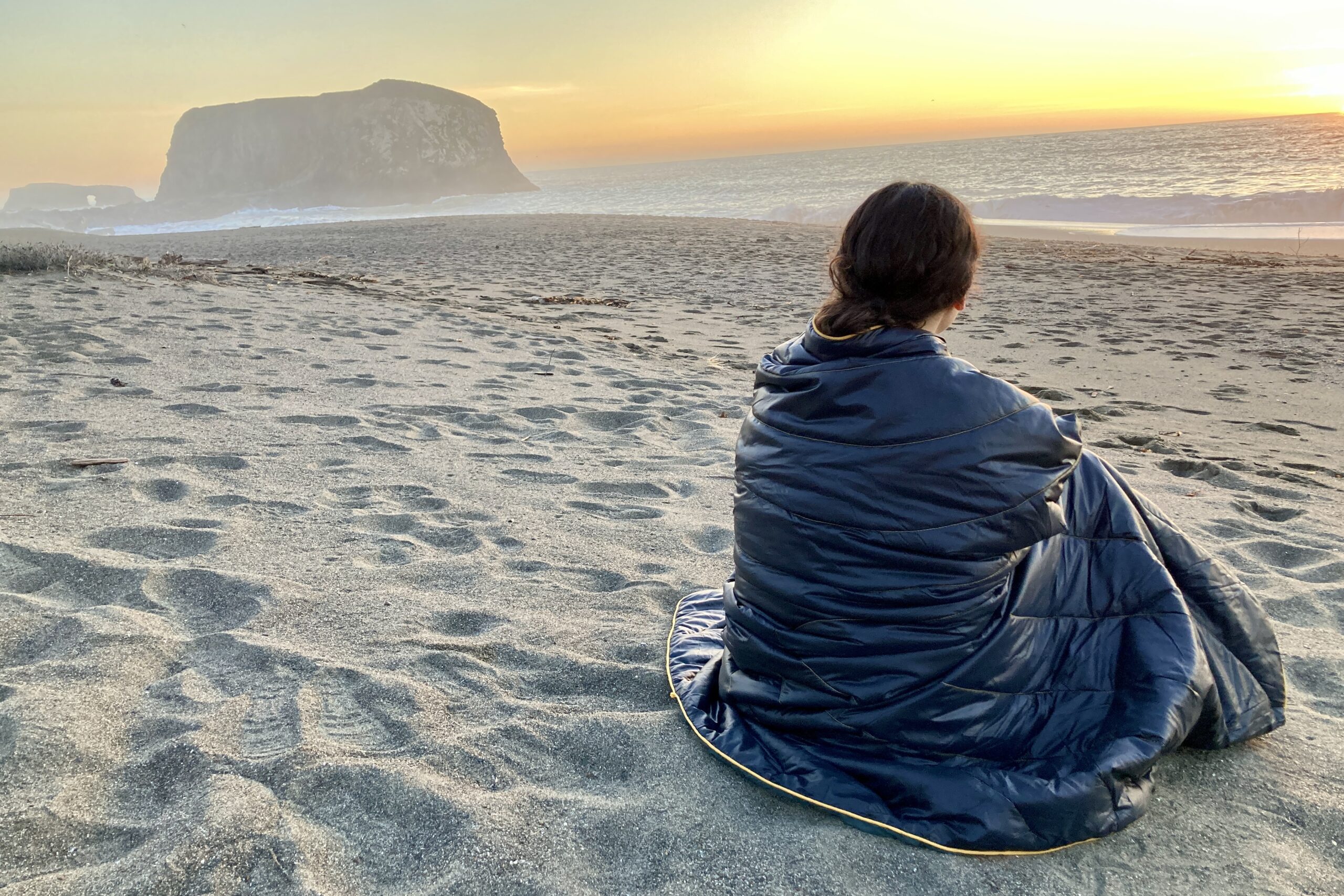 Person wrapped in the Rumpl Original Puffy Blanket while sitting on the beach at sunset.