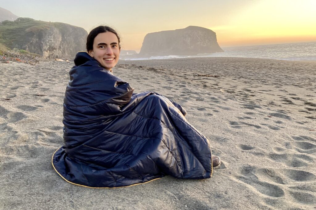 Women sitting on beach curled up in the Rumpl Original Puffy Blanket.