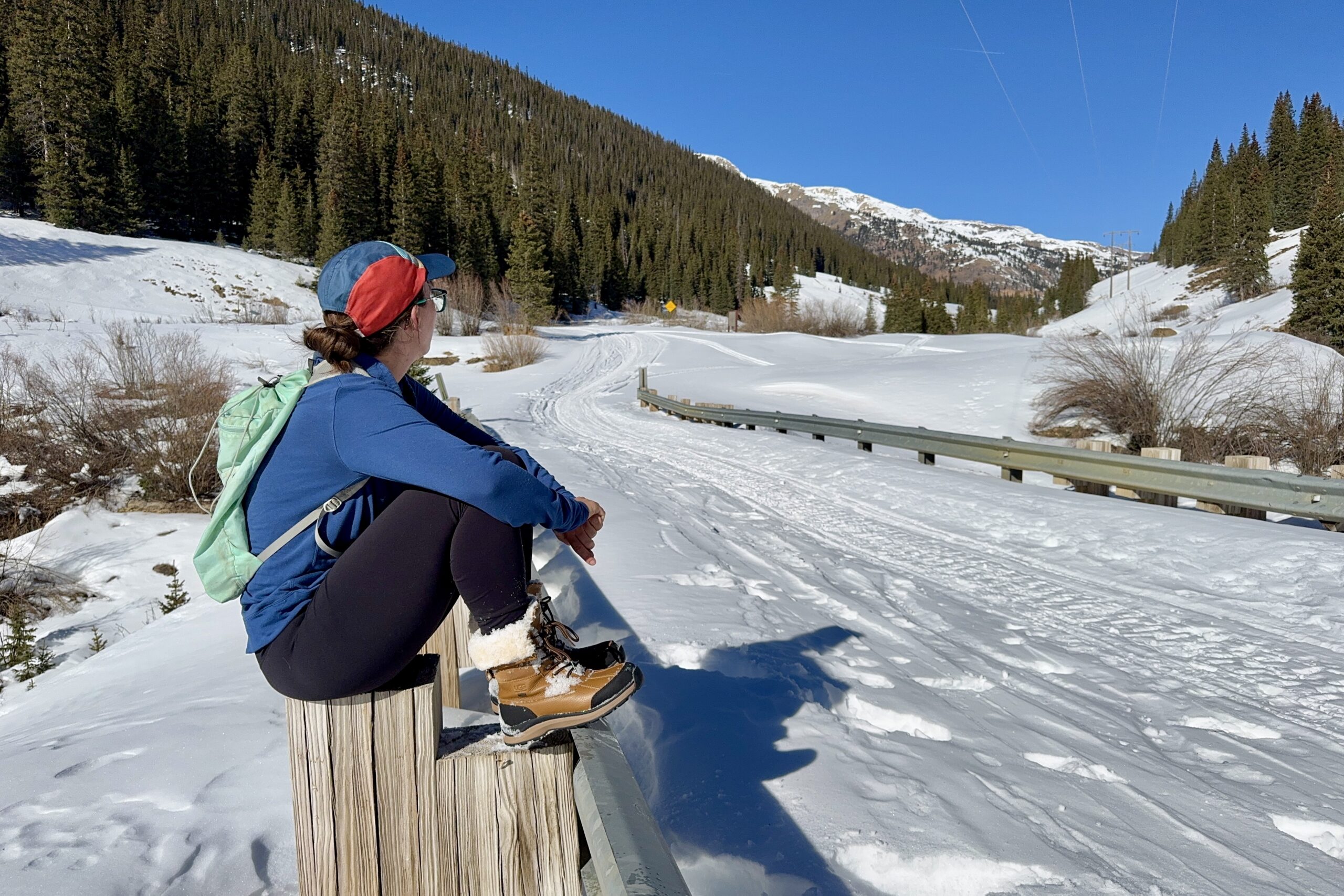 A person wearing the UGG boots is sitting on a rail looking at a mountain view.