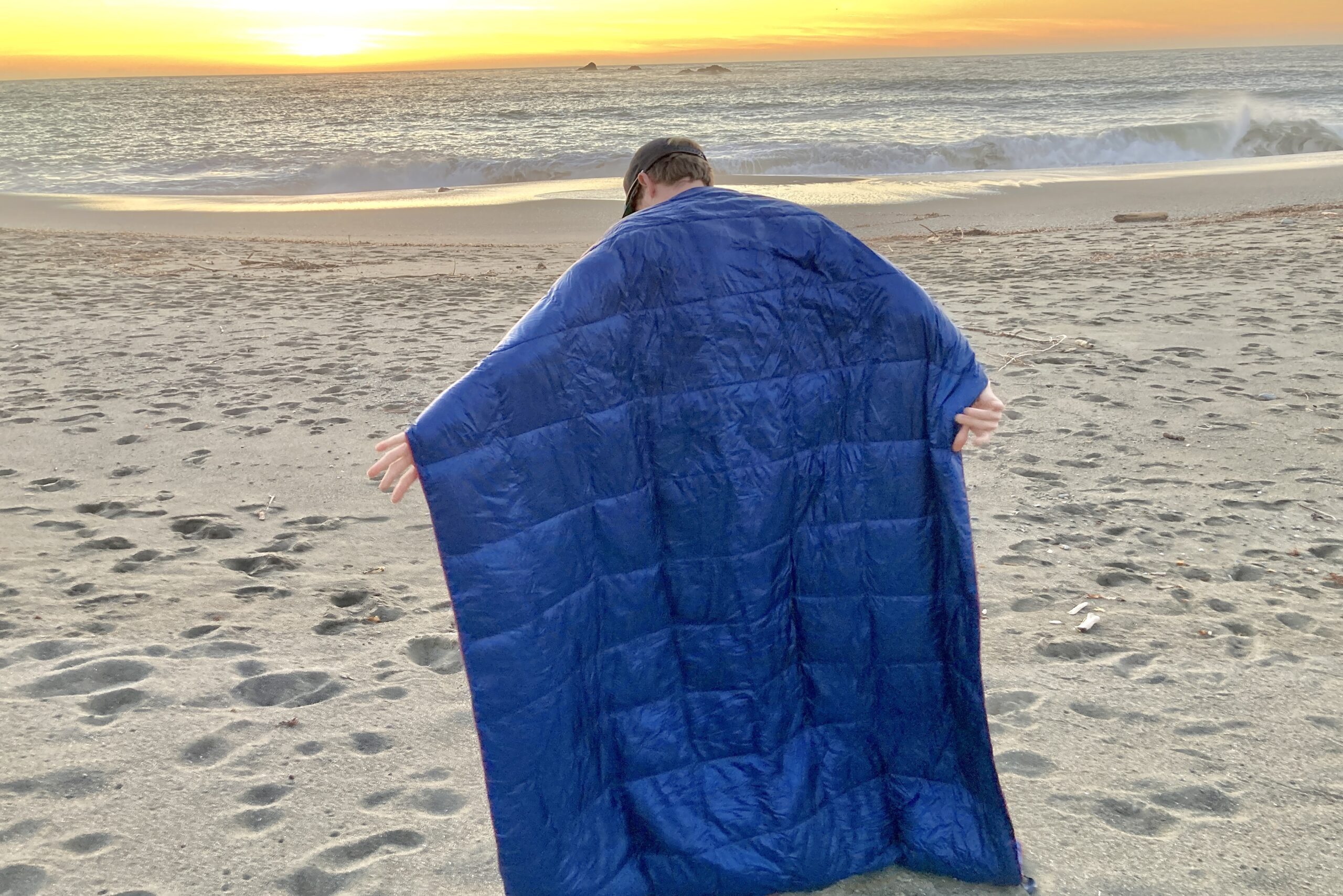 Person standing facing the sunset on the beach while wearing the Horizon Hound Trek Blanket