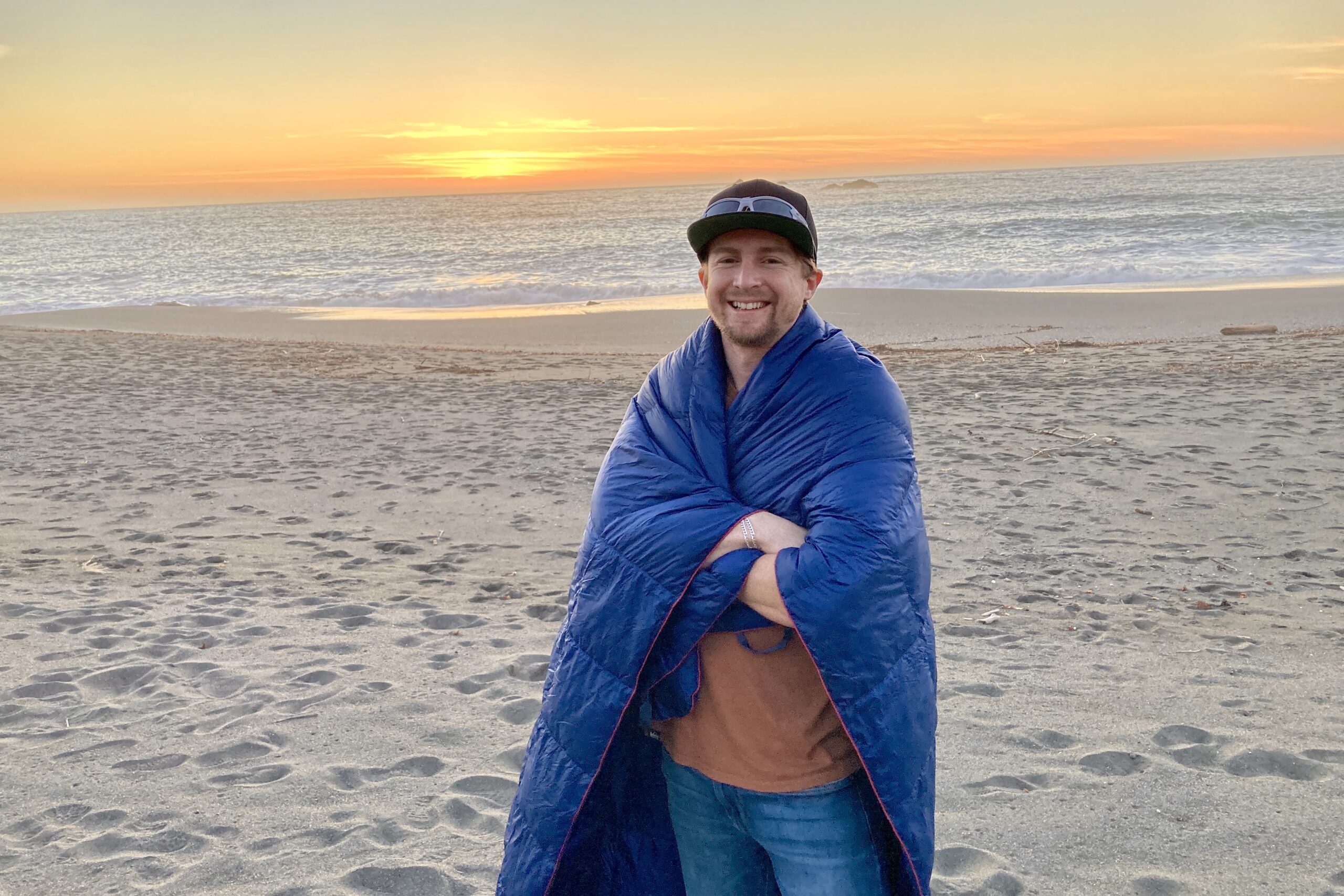 Man standing on the beach facing the camera with the Horizon Hound Trek Blanket wrapped around his shoulders.