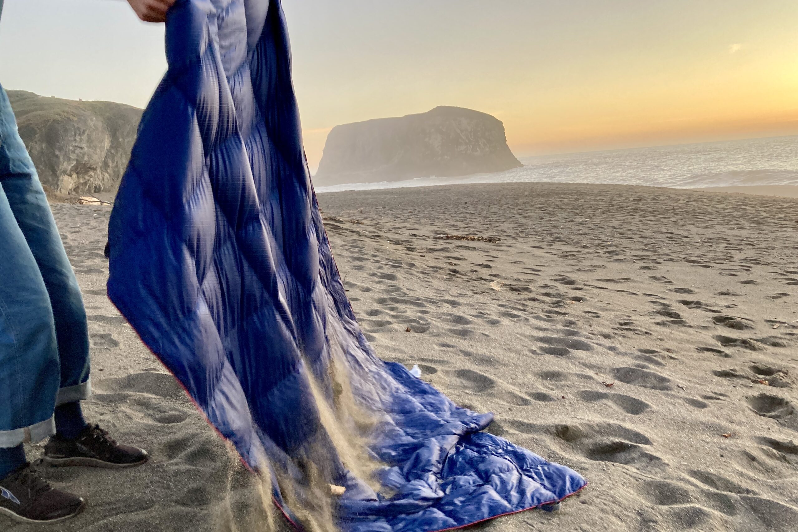 Person shaking sand off of the Horizon Hound Trek Blanket on the beach.