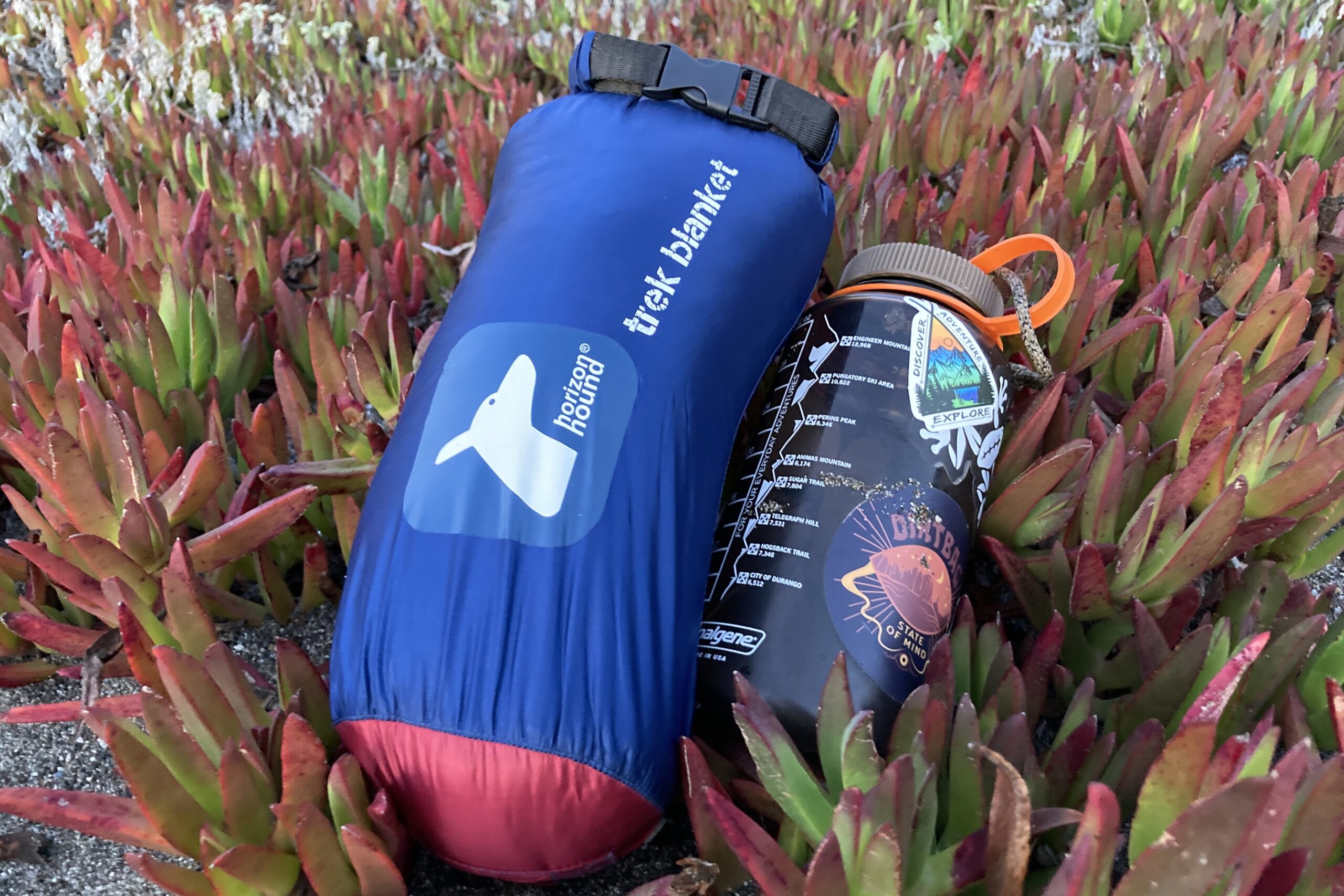 Laying the Horizon Hound Trek Blanket in its carry bag next to a Nalgene to show the relative size difference and packability of the blanket