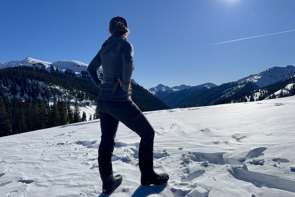 A person observing a mountain view while wearing the Neo Classic Tall boots in the snow.