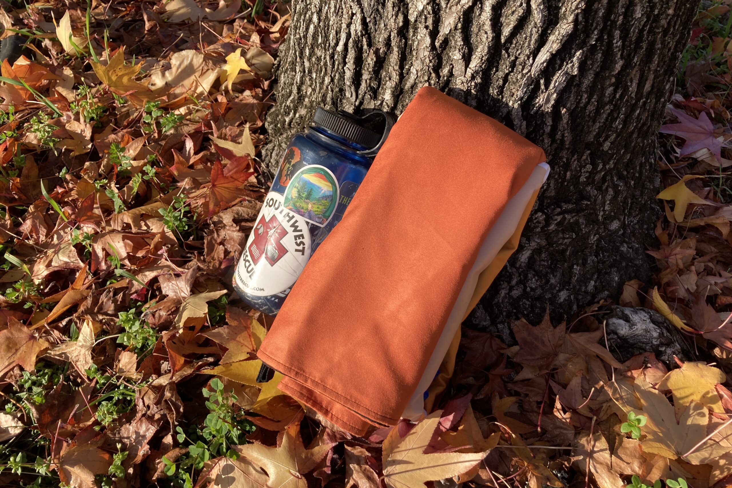 Close up showing the size of the Nomadix Festival Blanket when rolled up, sitting next to a 32 oz Nalgene by a tree trunk.