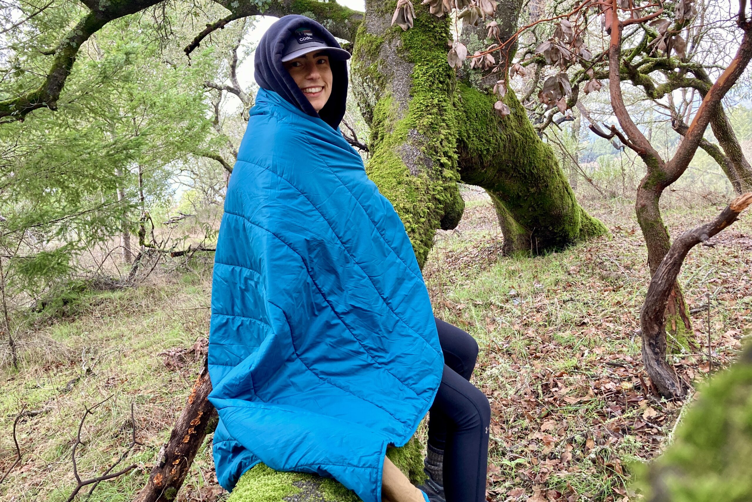 Person wrapped in the REI camp blanket while sitting on a mossy tree branch