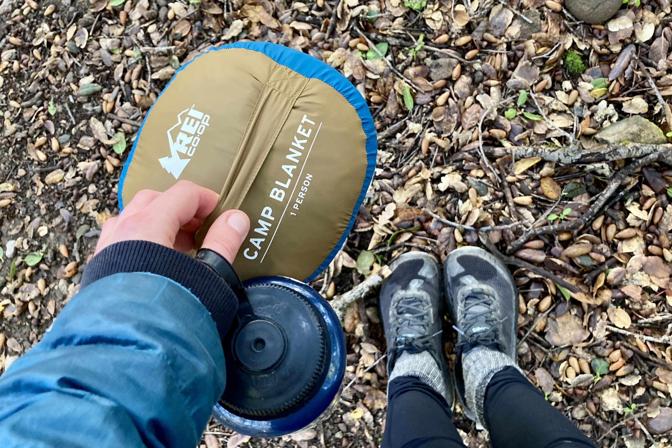 Close up showing someone carrying the REI Camp blanket by the strap on the bottom of the carry bag, while also holding a Nalgene.