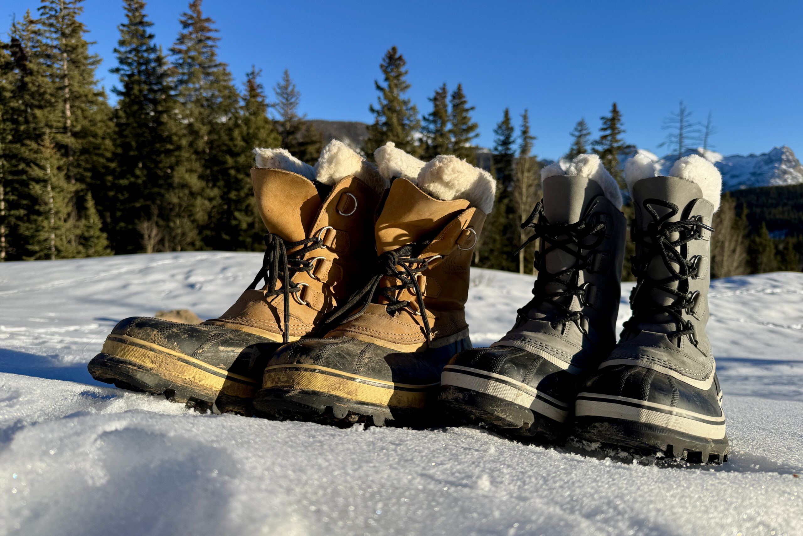 A pair of brown Sorels sits next to a pair of gray Sorels in the snow with woods in the background.