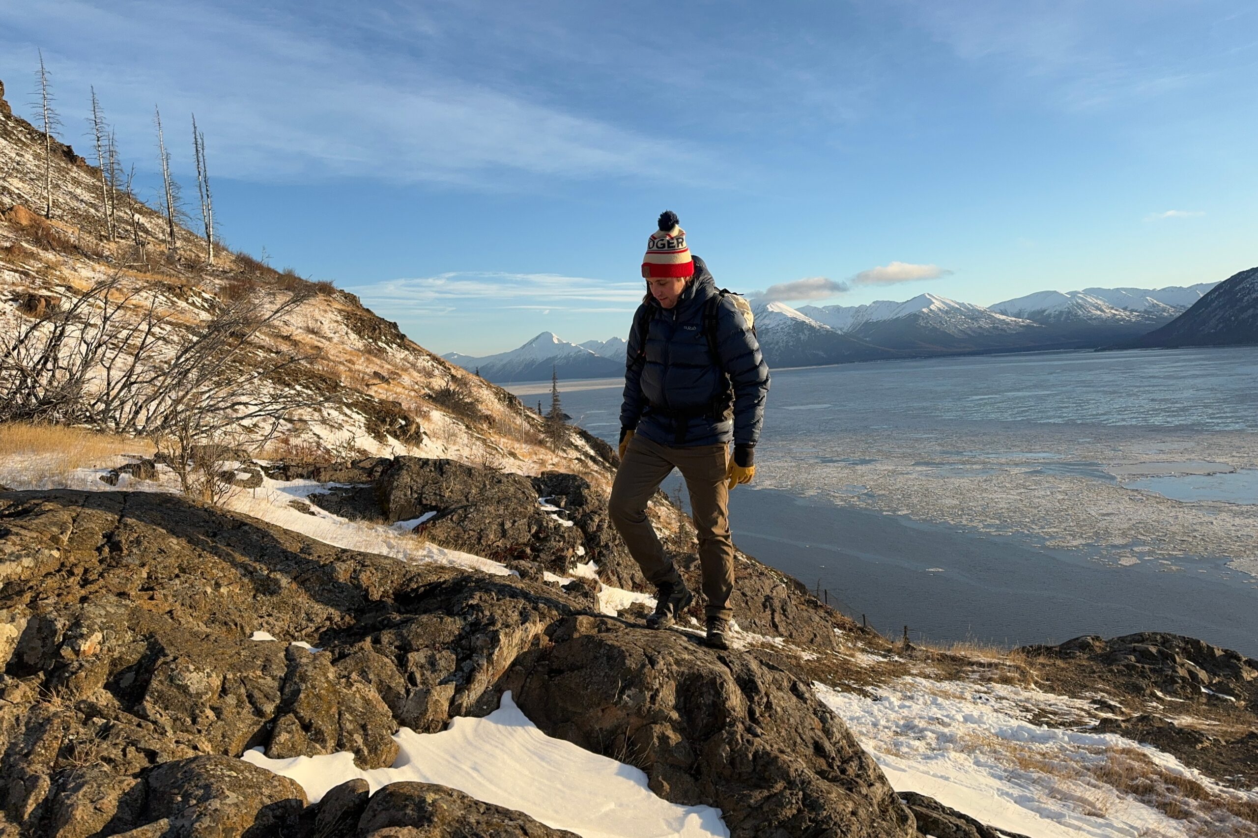 A man walks up a rocky ridge with ocean and mountains in the background.