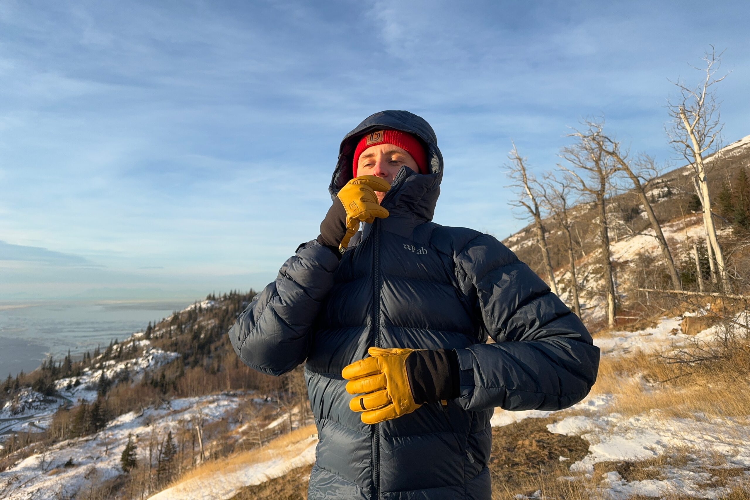 A man zips up a down jacket on a sunny mountainside.