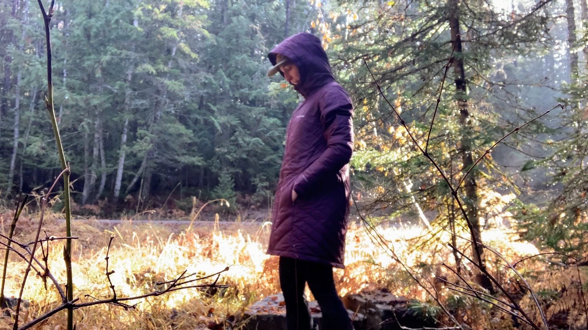 a woman walking in a forest next to a dirt road with sunlight peaking through behind her. She's wearing a columbia long hooded jacket with her hands in the pockets and the hood over her ball cap.