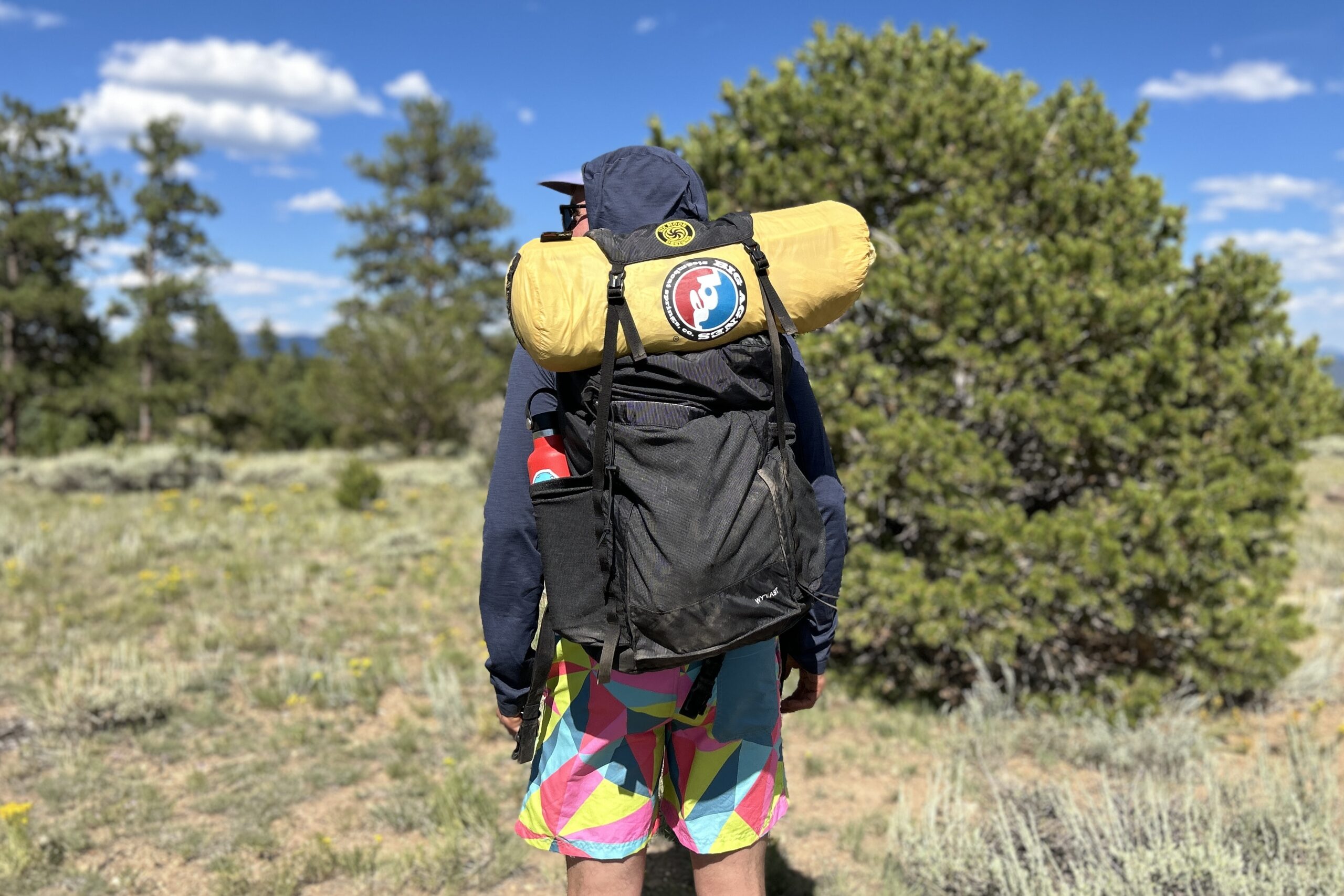 A man wearing a daypack with a large tent strapped to the top looks into the distance on a bluebird day in the forest.