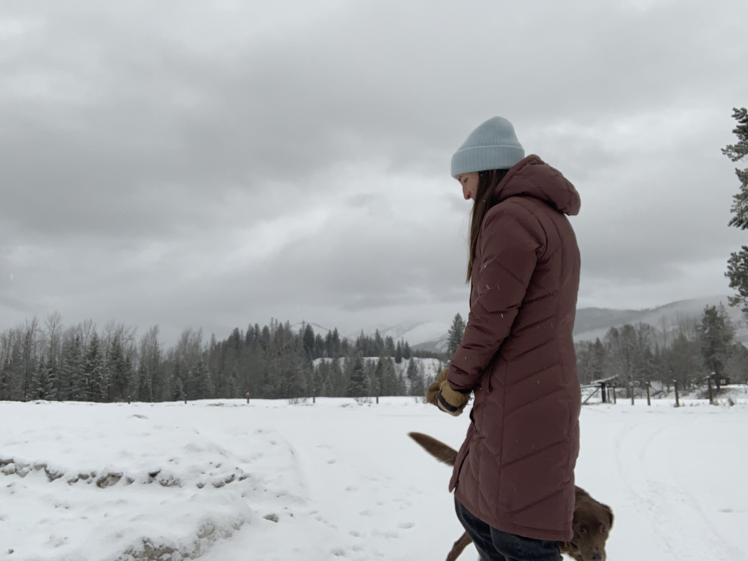 a side view of the baffle stitching on the patagonia down with it parka. A woman is walking along a snowy road with snow-covered foothills in the distance