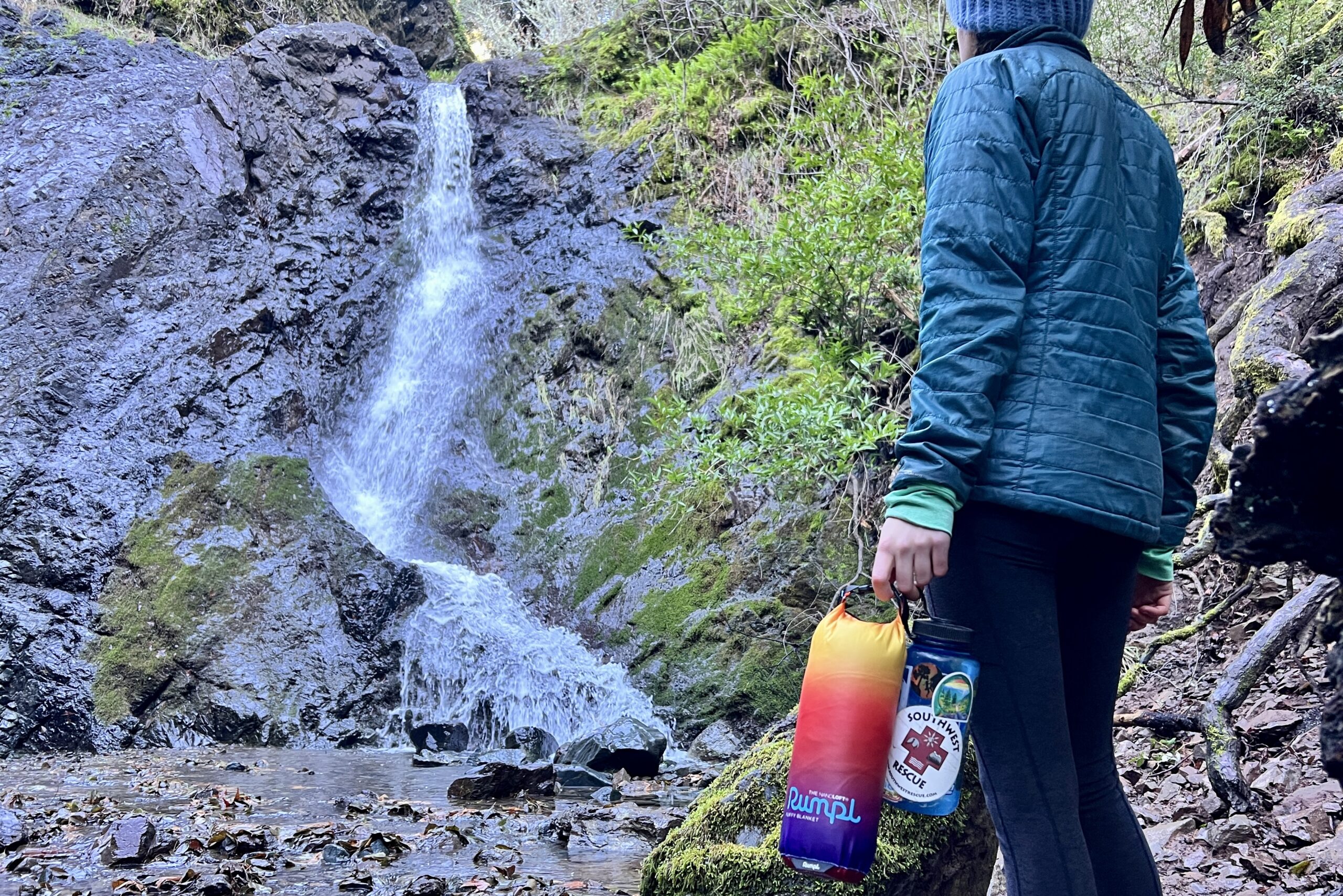 Person standing in front of a waterfall, holding both a Nalgene water bottle and the Rumpl NanoLoft Puffy Blanket, comparing the similar sizes.