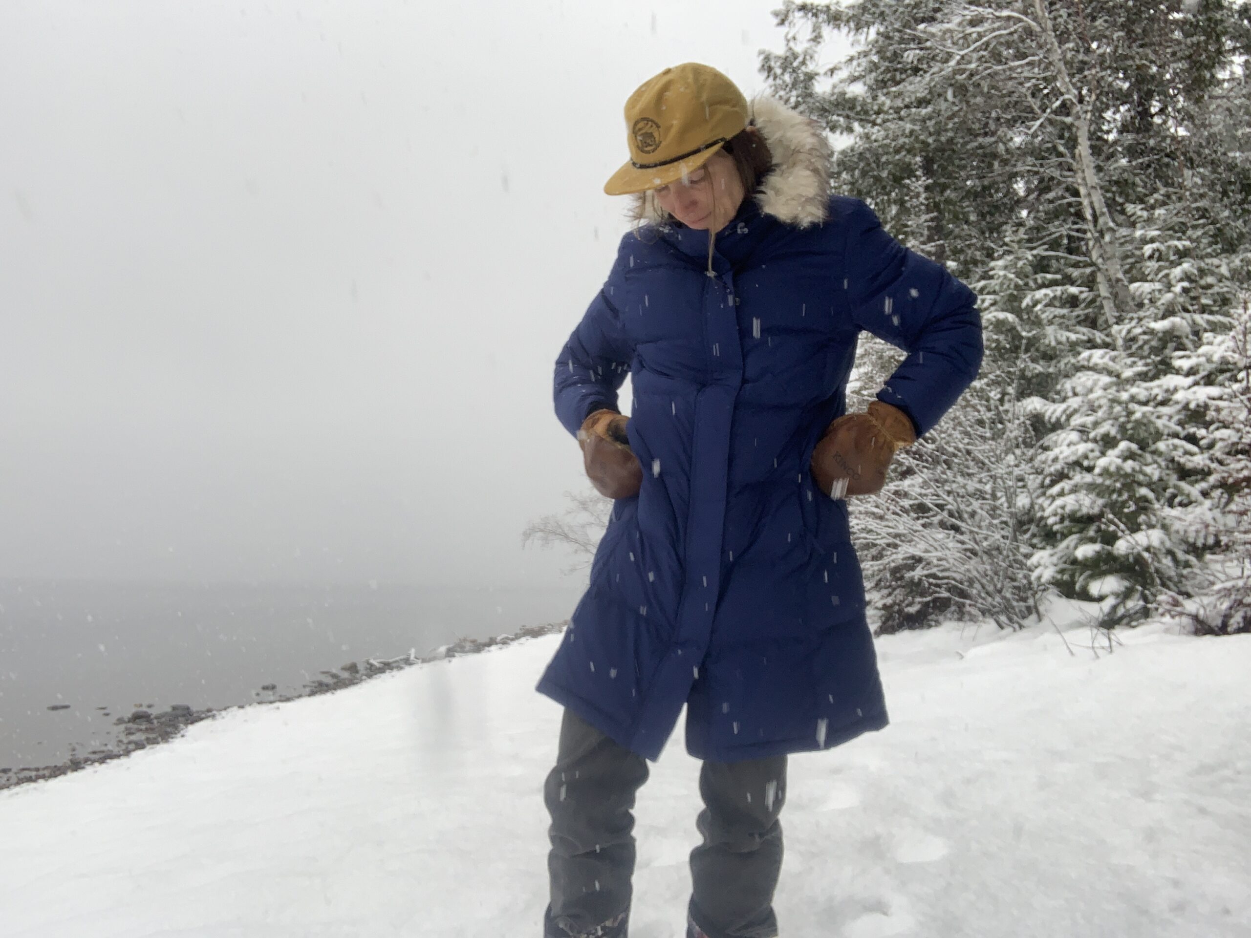 a woman standing next to a lake while it is snowing. She is wearing an llbean ultrawarm down puffy parka