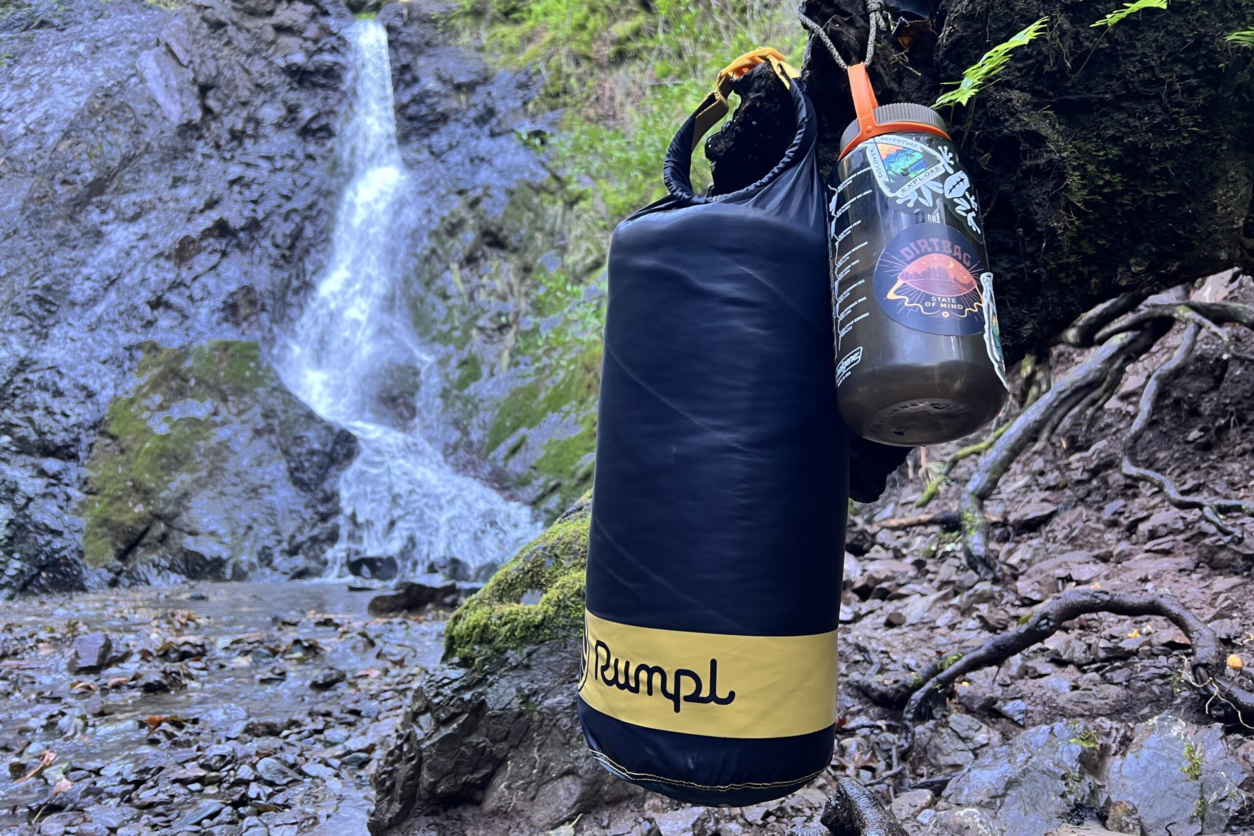 The Rumpl Original Puffy Blanket hangs from a tree in its carry case next to a Nalgene with a waterfall in the background.