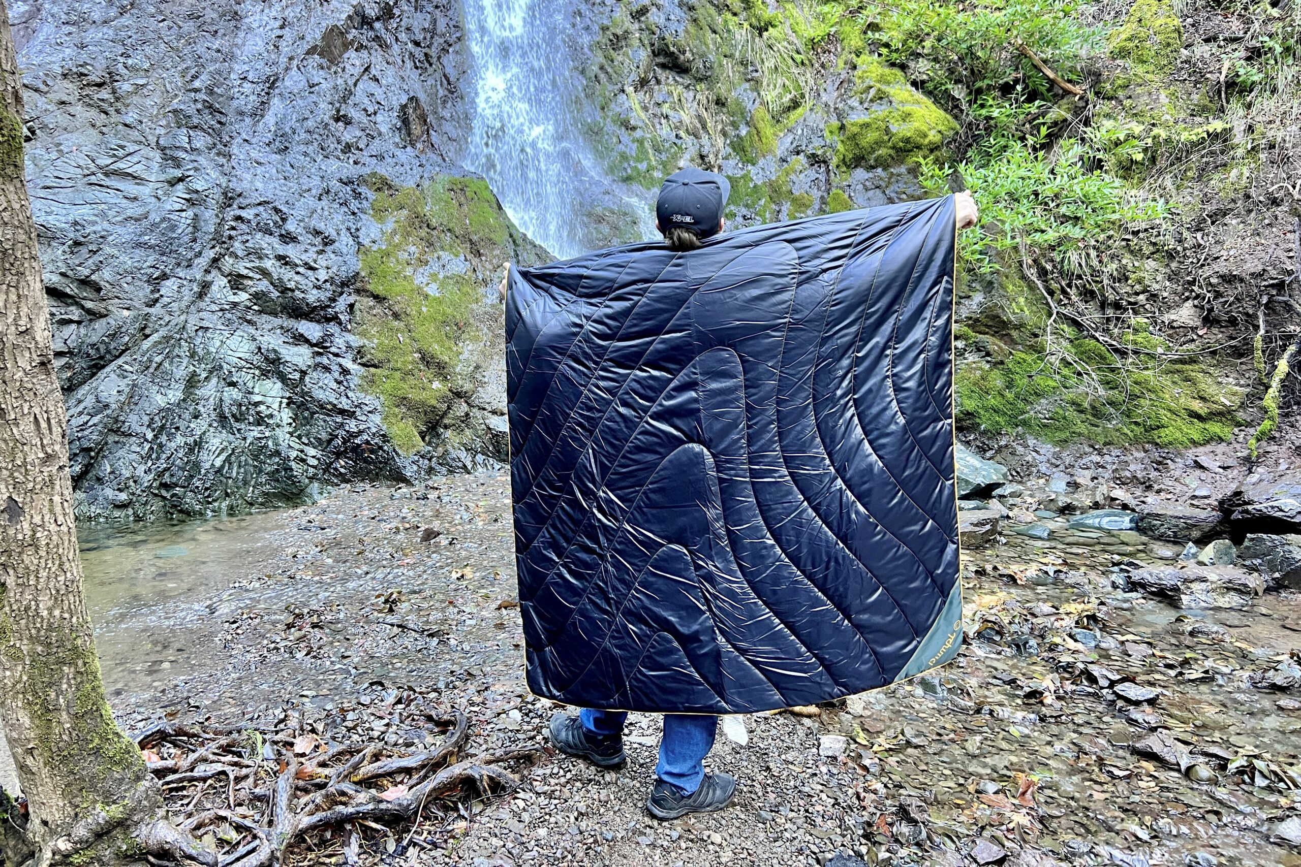 Man standing before a waterfall with the Rumpl Original Puffy Blanket around his back.