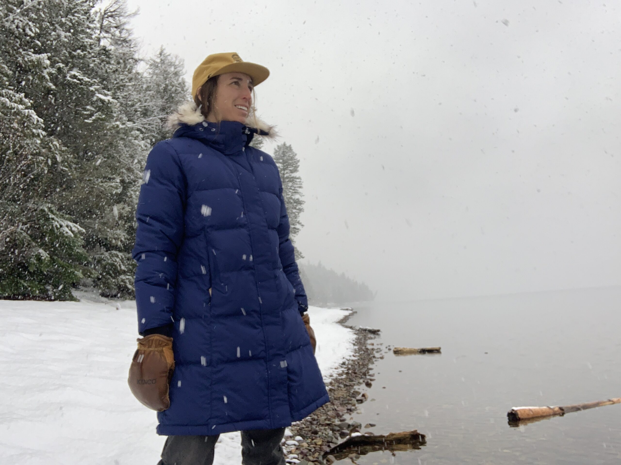 a woman looks out over a lake while it is snowing. She is warm and cozy testing the quality of the llbean ultrawarm down parka