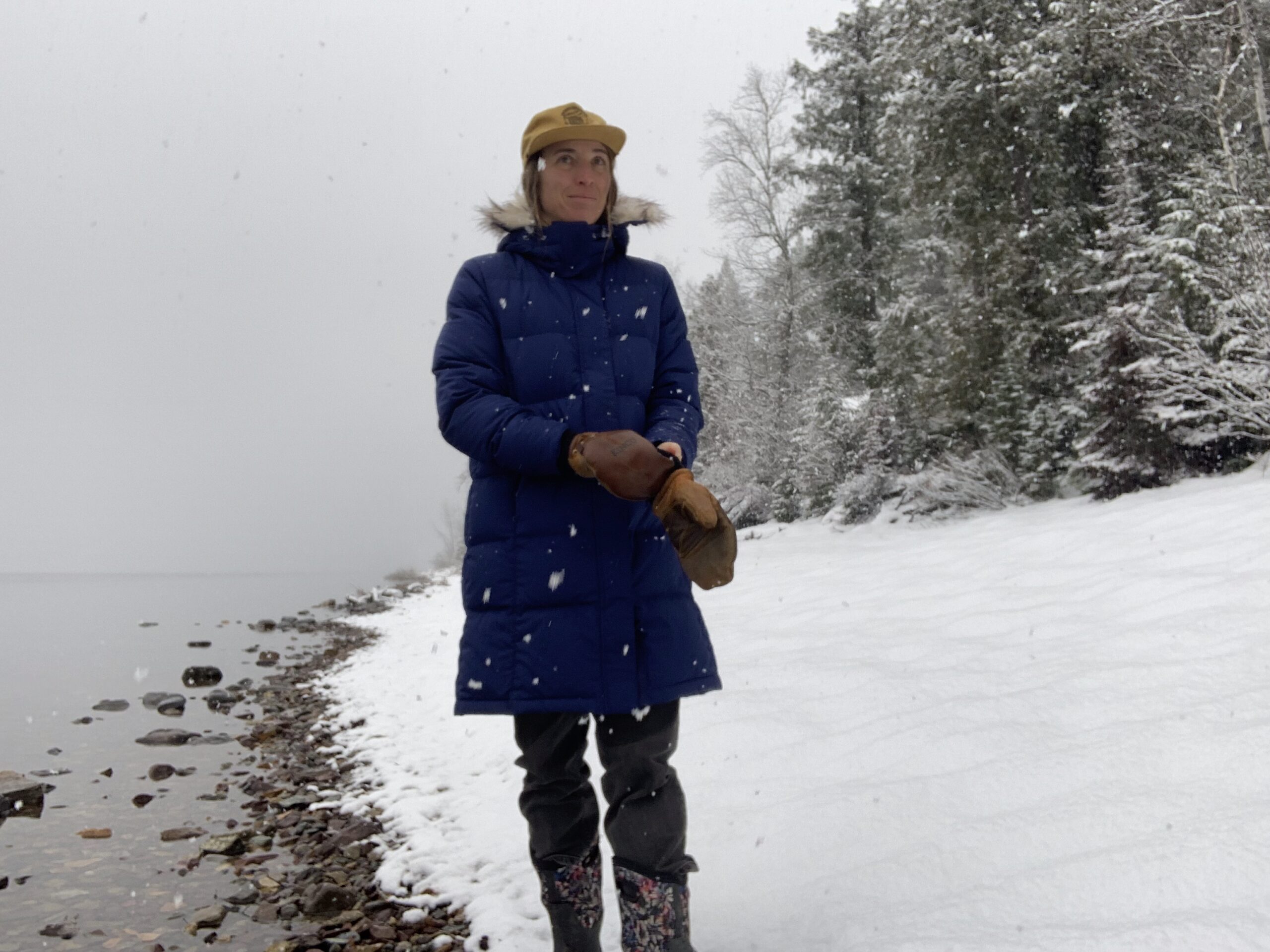 a woman standing by a lake while it is snowing puts on her mittens while wearing the ll bean ultrawarm 3/4 down parka