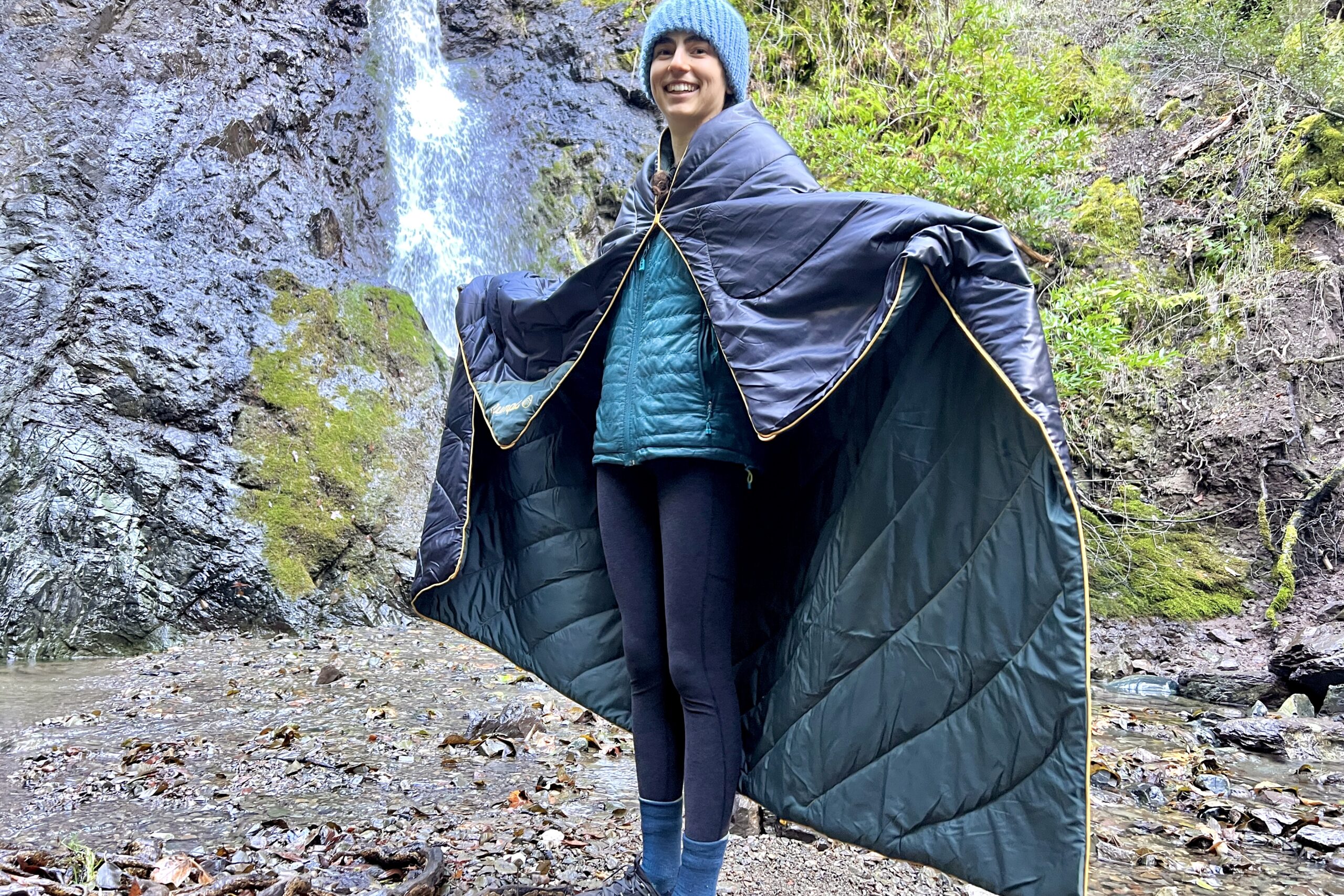 Woman standing in front of a waterfall holding out the corners of the Rumpl Original Puffy Blanket as a cape.