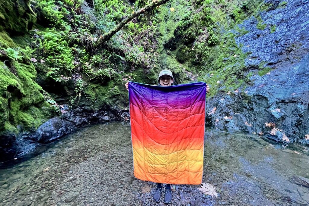 Person holding up the Rumpl NanoLoft Puffy Blanket with a mossy wooded background, to compare blanket size to the person’s height.