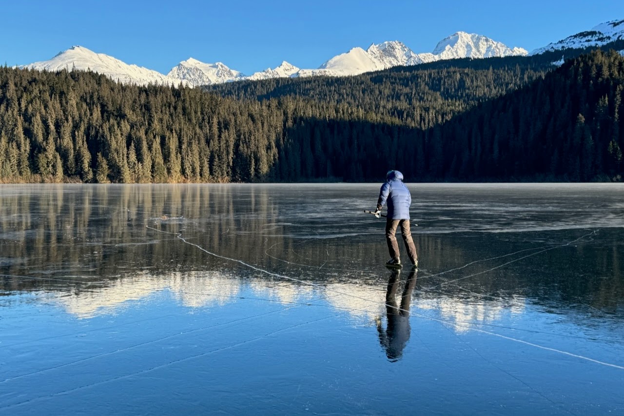 A man ice skates away from the camera with mountain views.