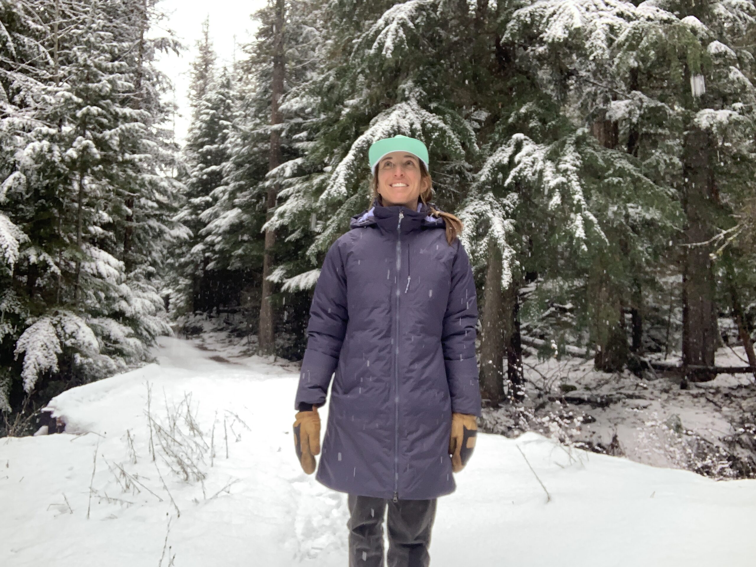 a woman standing in front of a snowy bridge in a forest looking at the snowfalling