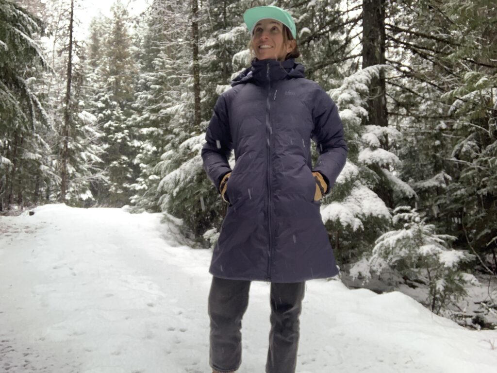 a woman in a cedar forest while it is snowing. She is wearing the REI women's stormheng 850 hybrid parka