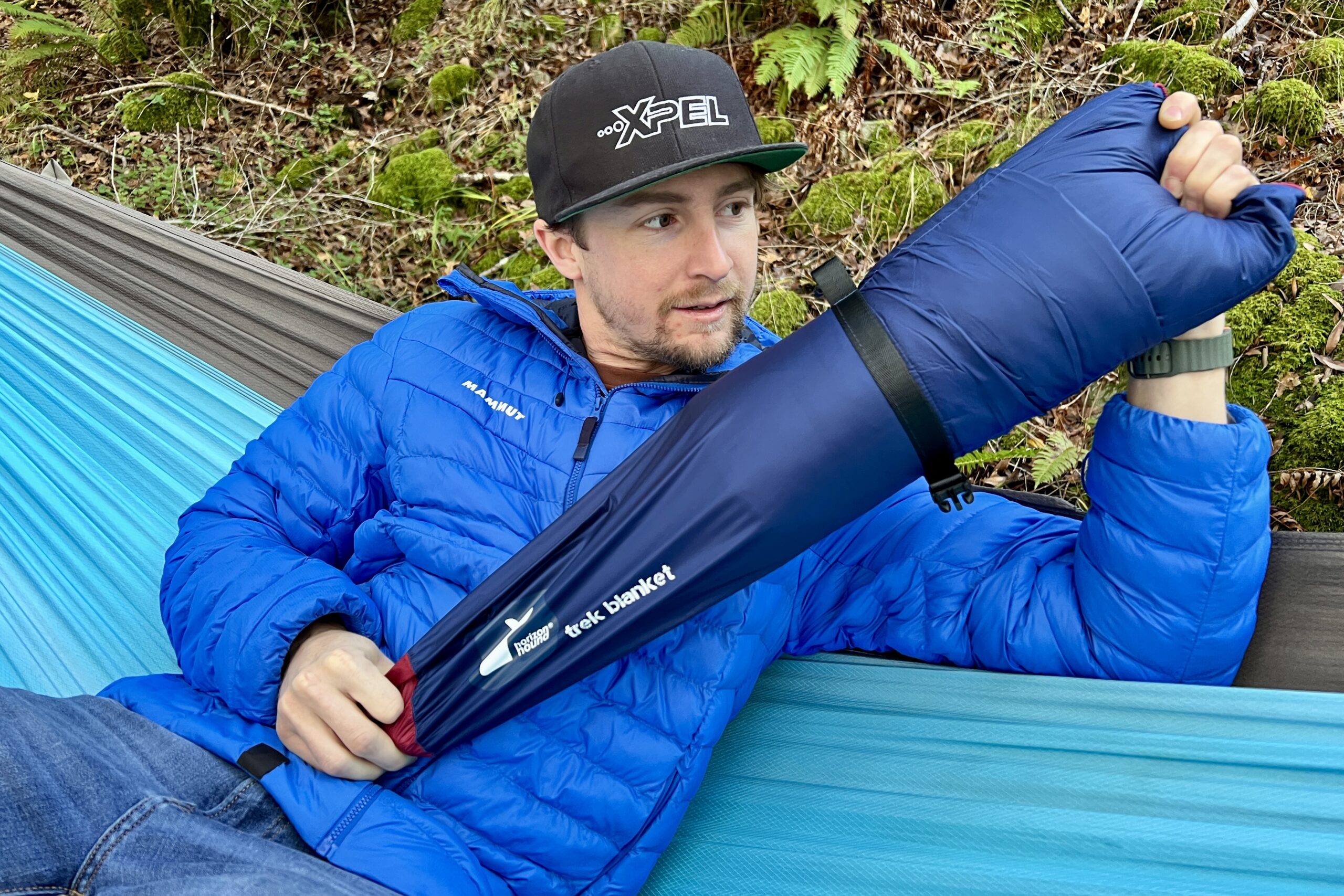 Man sitting in a hammock, pulling the Horizon Hound Trek Blanket out of its carry case.