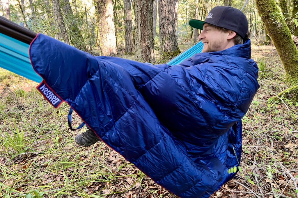 Showing a man sitting in a hammock wrapped in the Horizon Hound Trek Blanket.