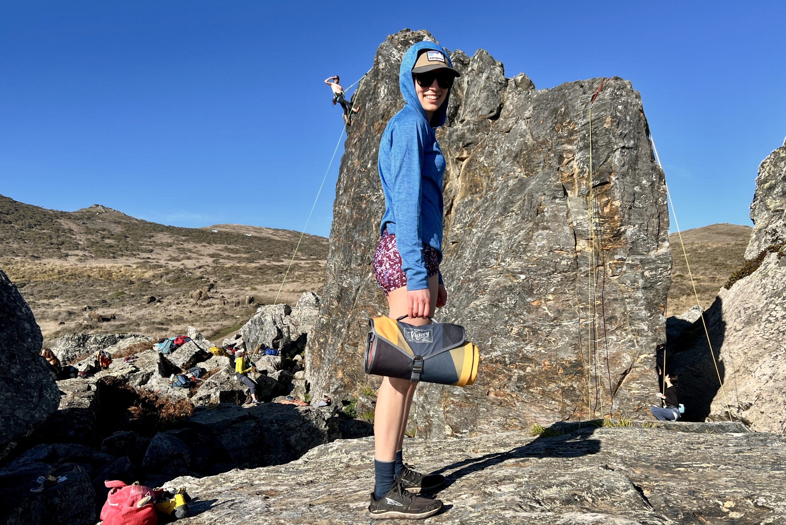 Woman carrying the Nemo Victory Patio Blanket by the handle, as it is rolled up in its carry case. She is staying in front of a rock climbing scene.