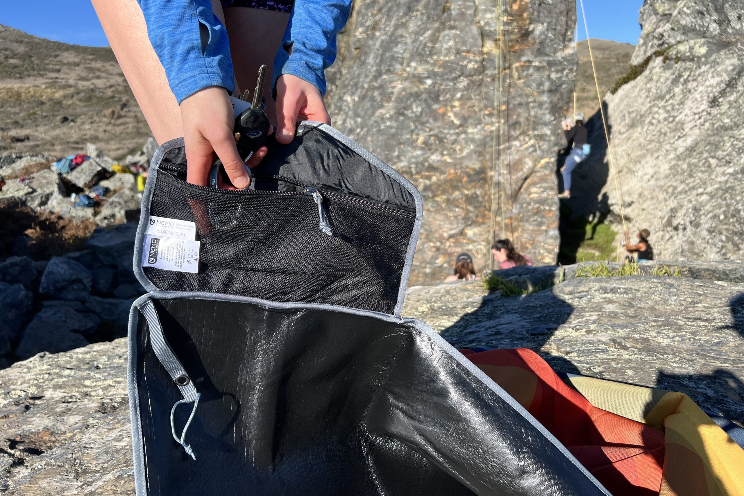 Holding up the Nemo Victory Patio Blanket to show its waterproof bottom layer and its included zippered pocket. Rock climbers are climbing in the background.