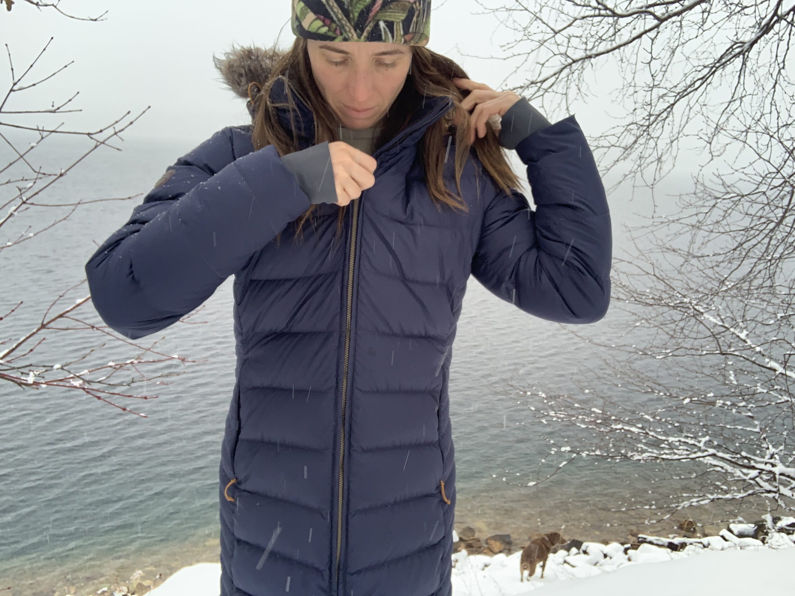 a woman standing in front of a lake zipping up the rab deep cover down parka