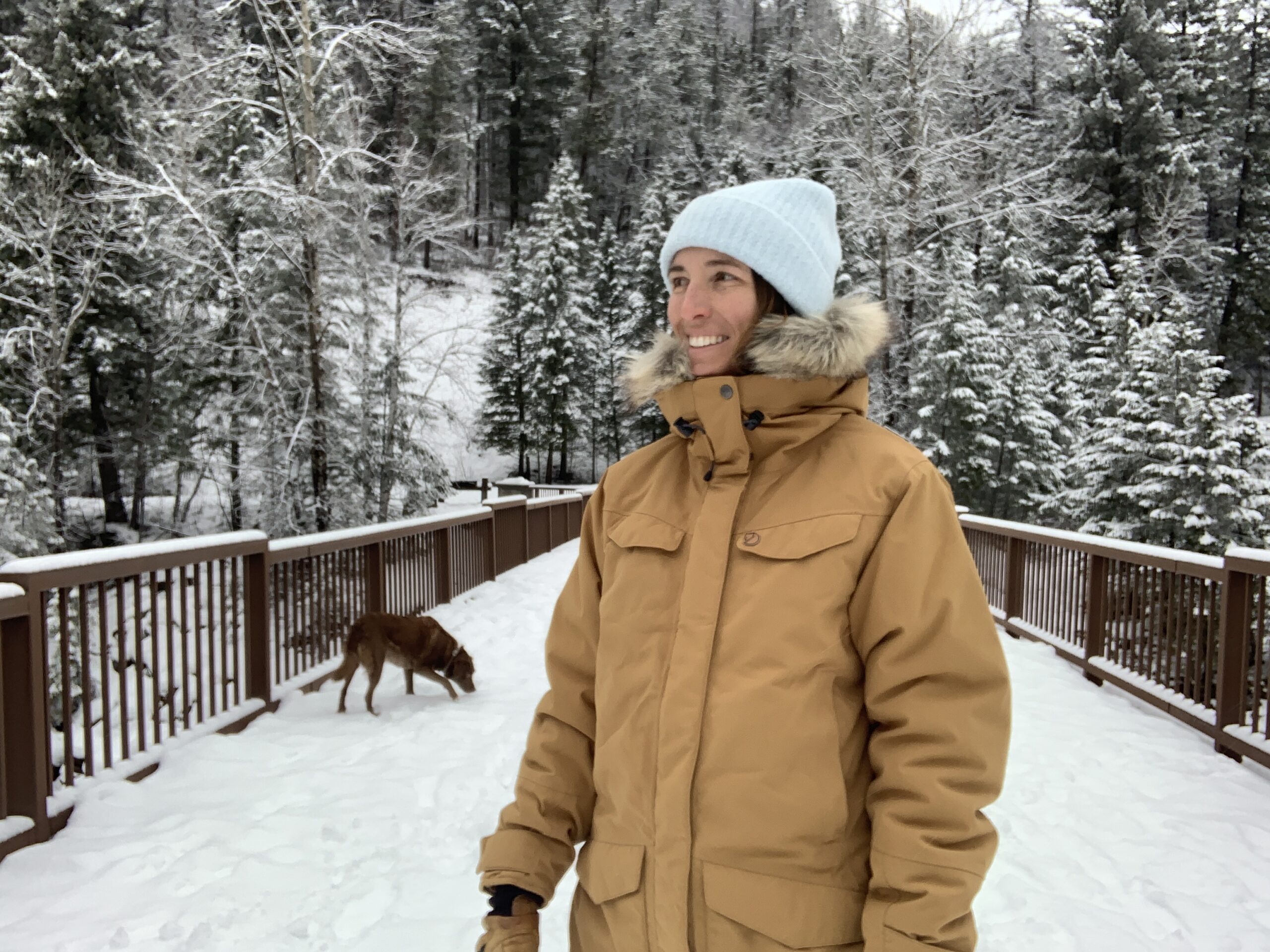 a woman smiles on a bridge over a glacial fed river in west glacier, MT. She's testing the quality and comfort and warmth of the fjallraven nuuk