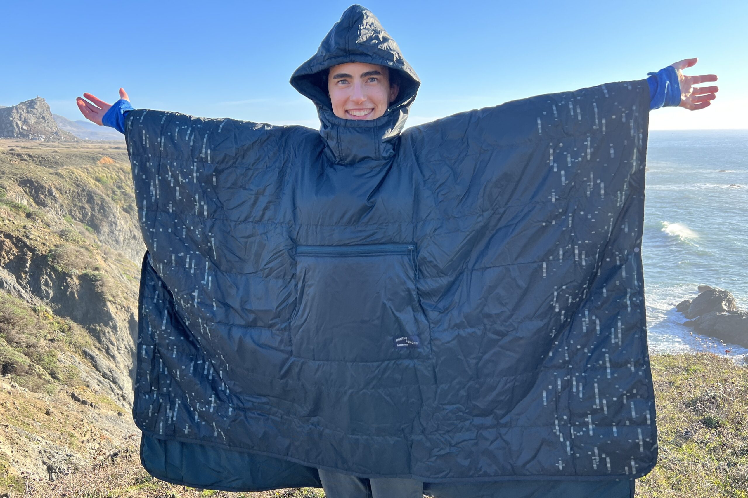 Woman standing with arms outstretched wearing the Therm-a-Rest Honcho Poncho on a coastal bluff.