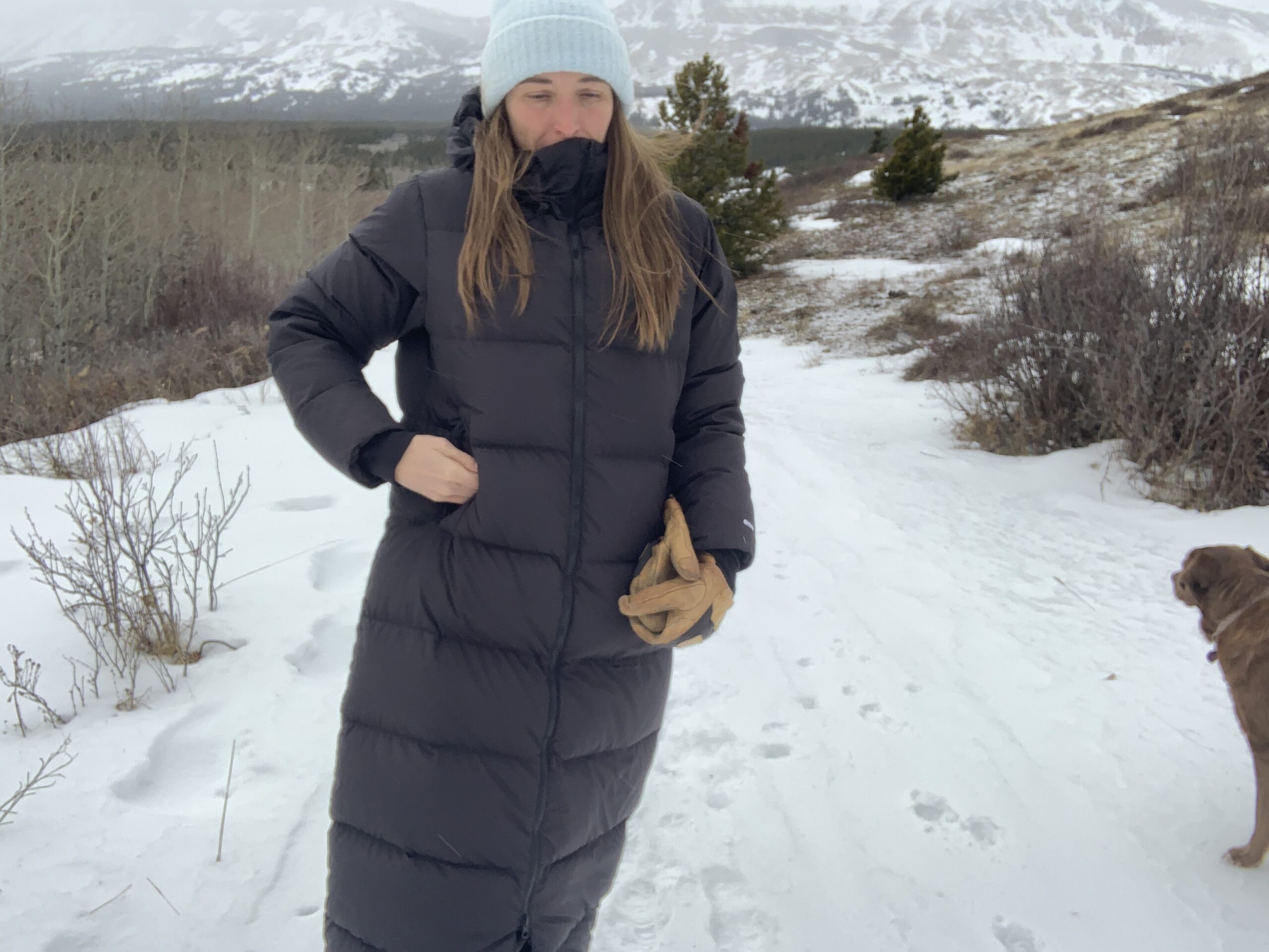 a woman puts her hands in her pocket while walking on a snowy trail with mountains in the distance.