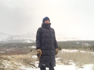 a woman stands in a windy meadow dotted with snow with mountains in the distance while staying warm in the outdoor research coze down parka