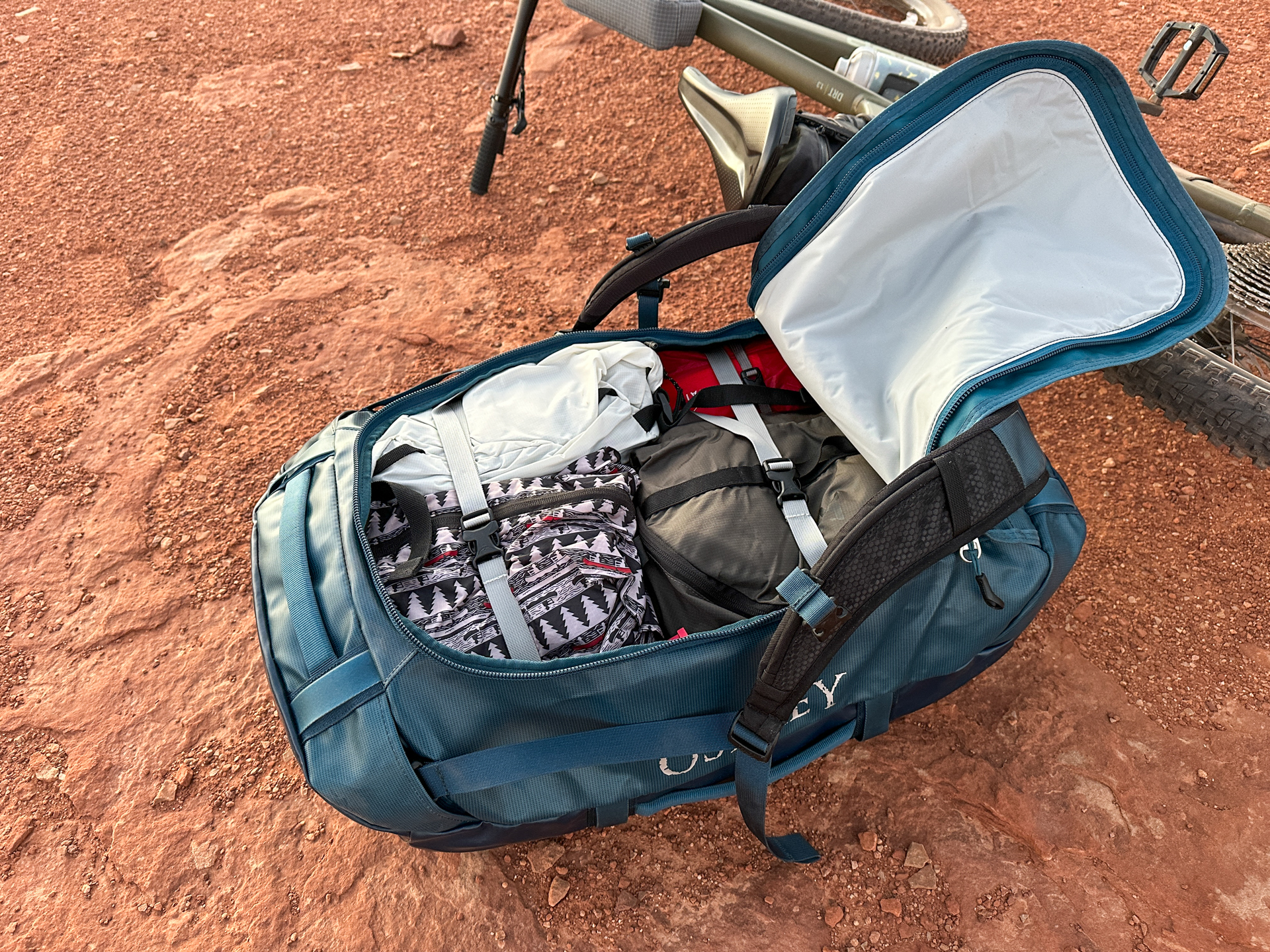 The Osprey Transporter sitting next to a bike in the desert open and packed full of clothing and gear.