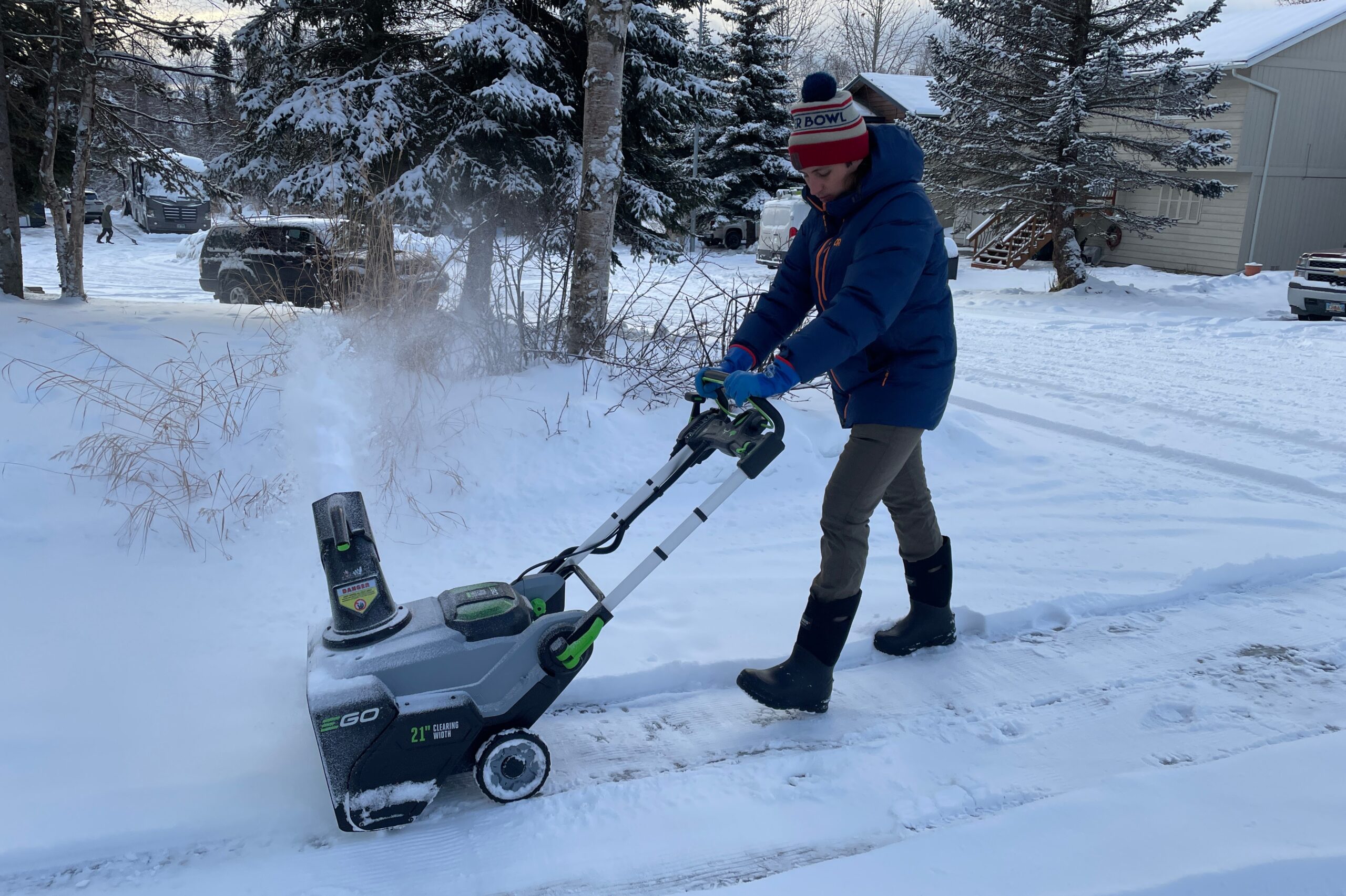 A man snowblows his driveway wearing a blue jacket.