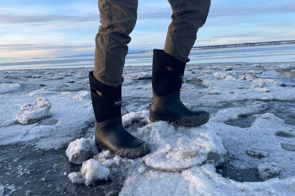 A man stands wearing boots in ice and mud.