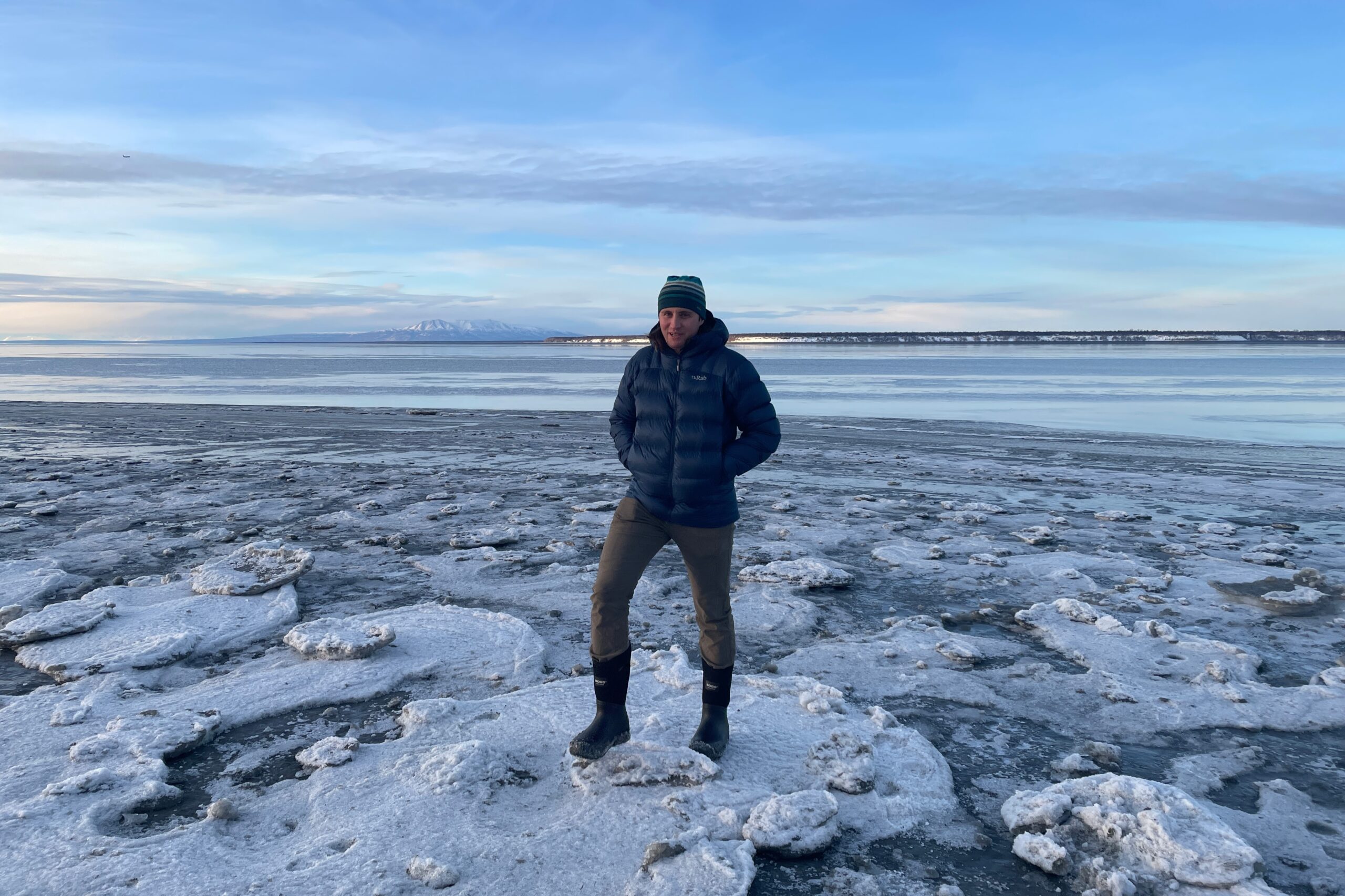 A man stands on a chunk of ice along a frozen coastline.