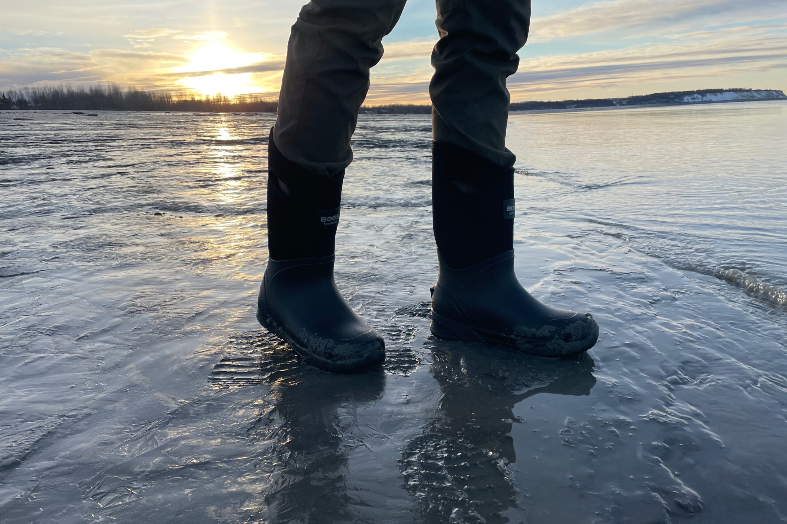 A man stands on a muddy shoreline with the sun setting in the background.