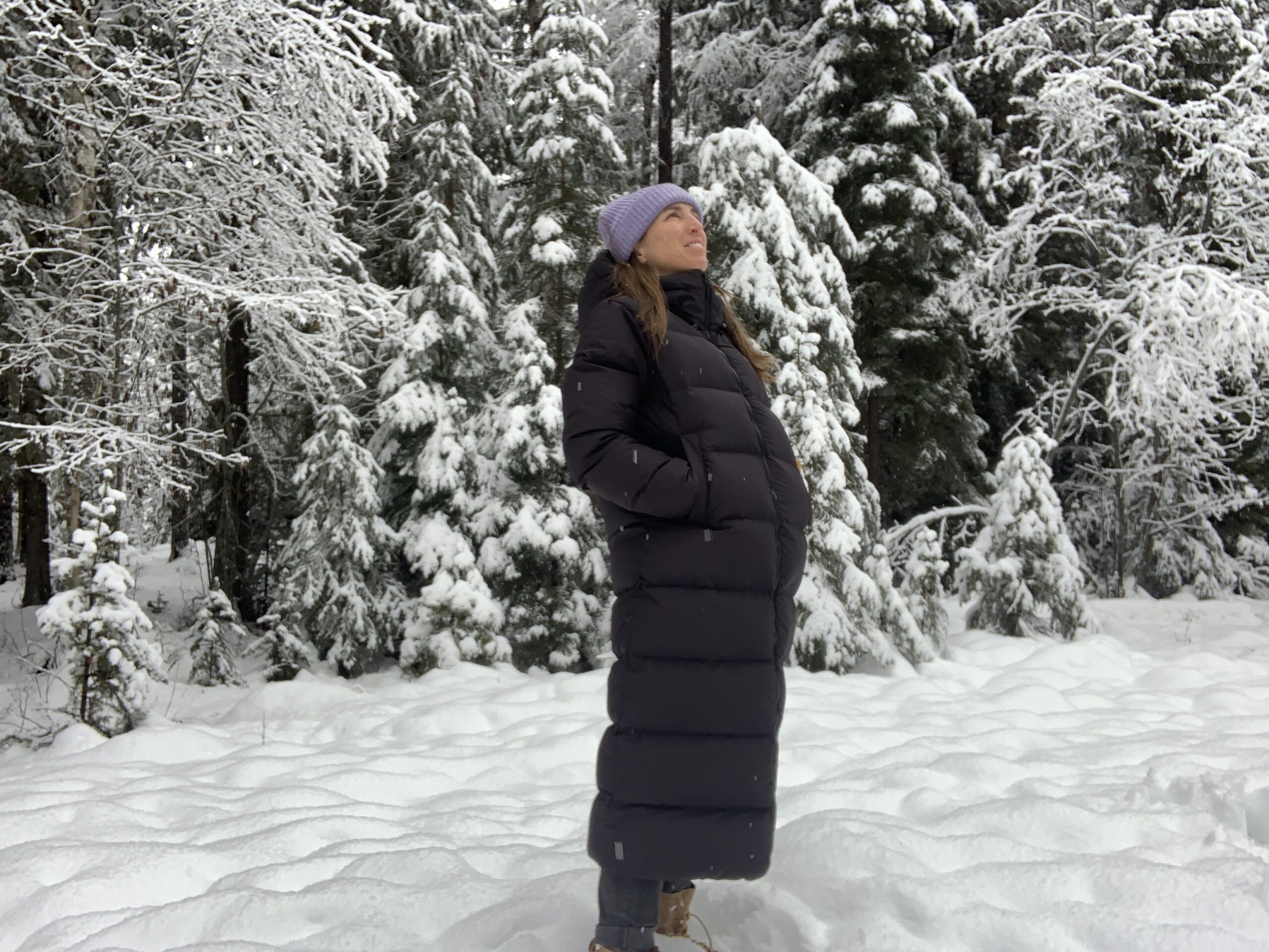 a woman stands in a snowy meadow looking up at the snow falling while wearing a long down parka covering her to the calves.