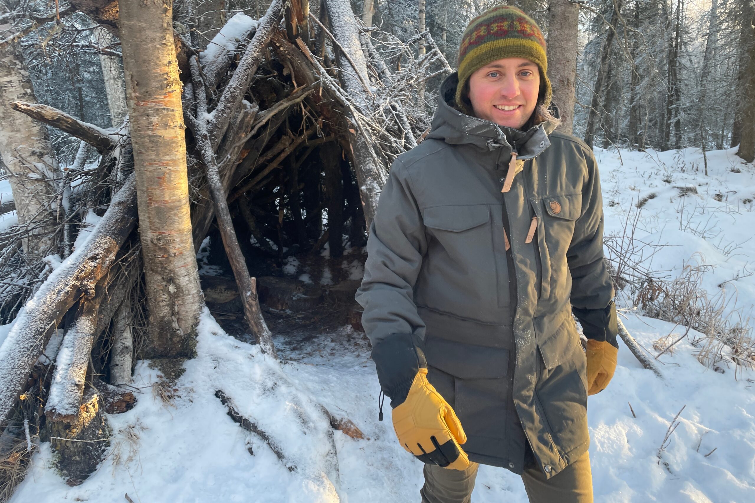 A man walks out of a shelter made of sticks in a snowy forest