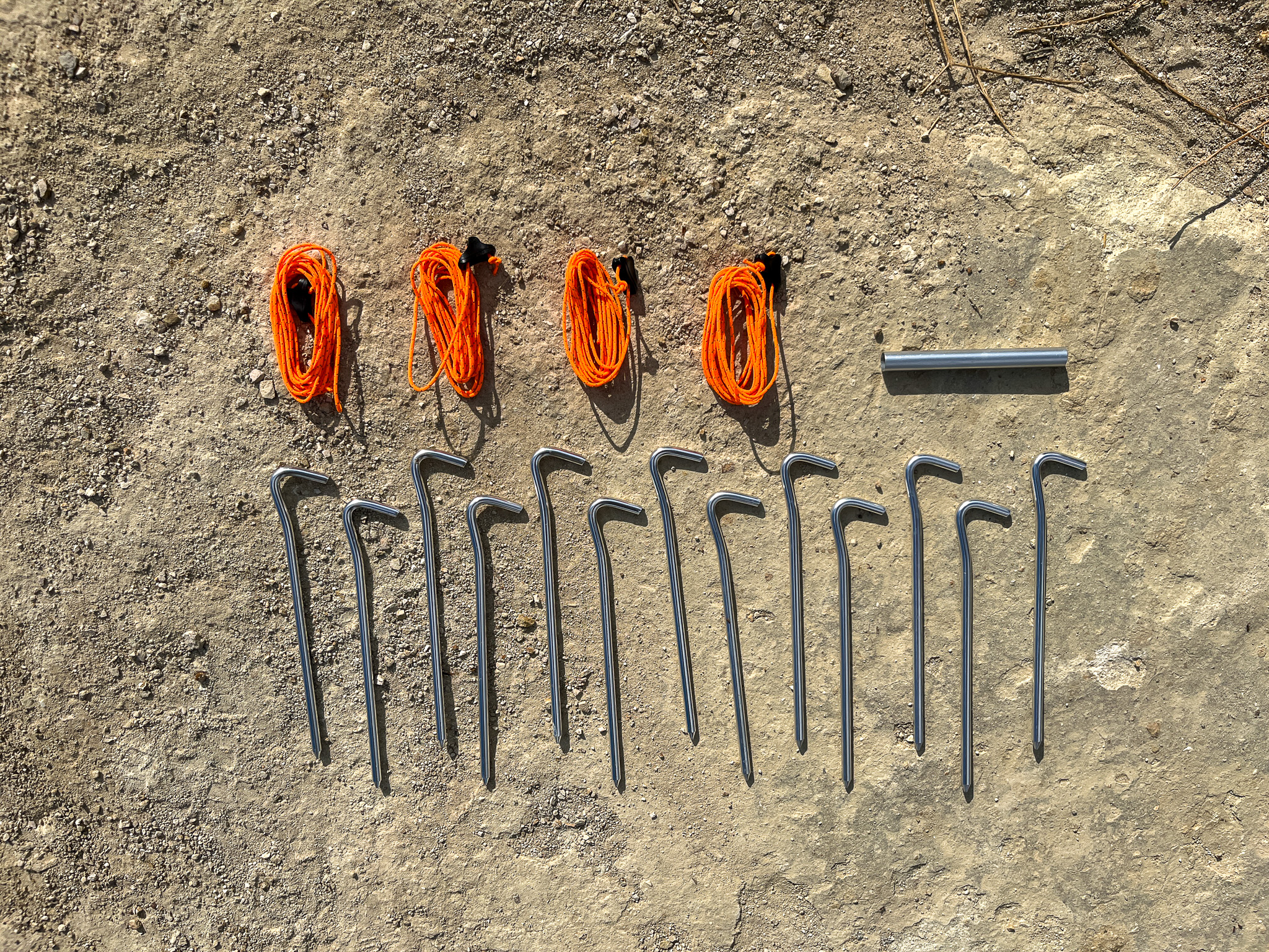 Closeup shot of the four included guy lines, pole repair splint, and hook-style stakes arranged neatly on the dirt ground.