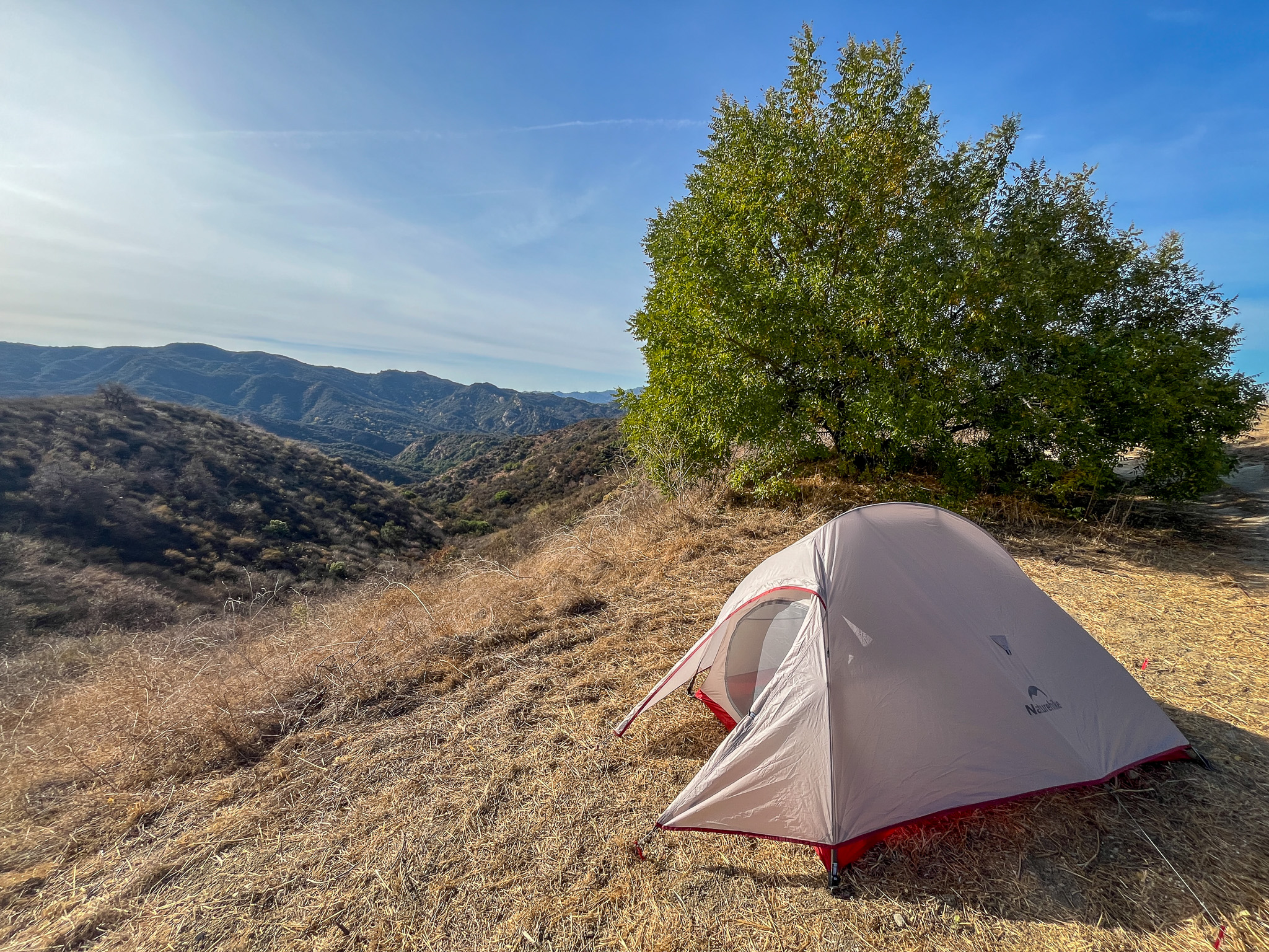 The Naturehike CloudUp 2 pitched on a scenic mountaintop campsite, overlooking rolling hills and a clear blue sky with a tree behind it.